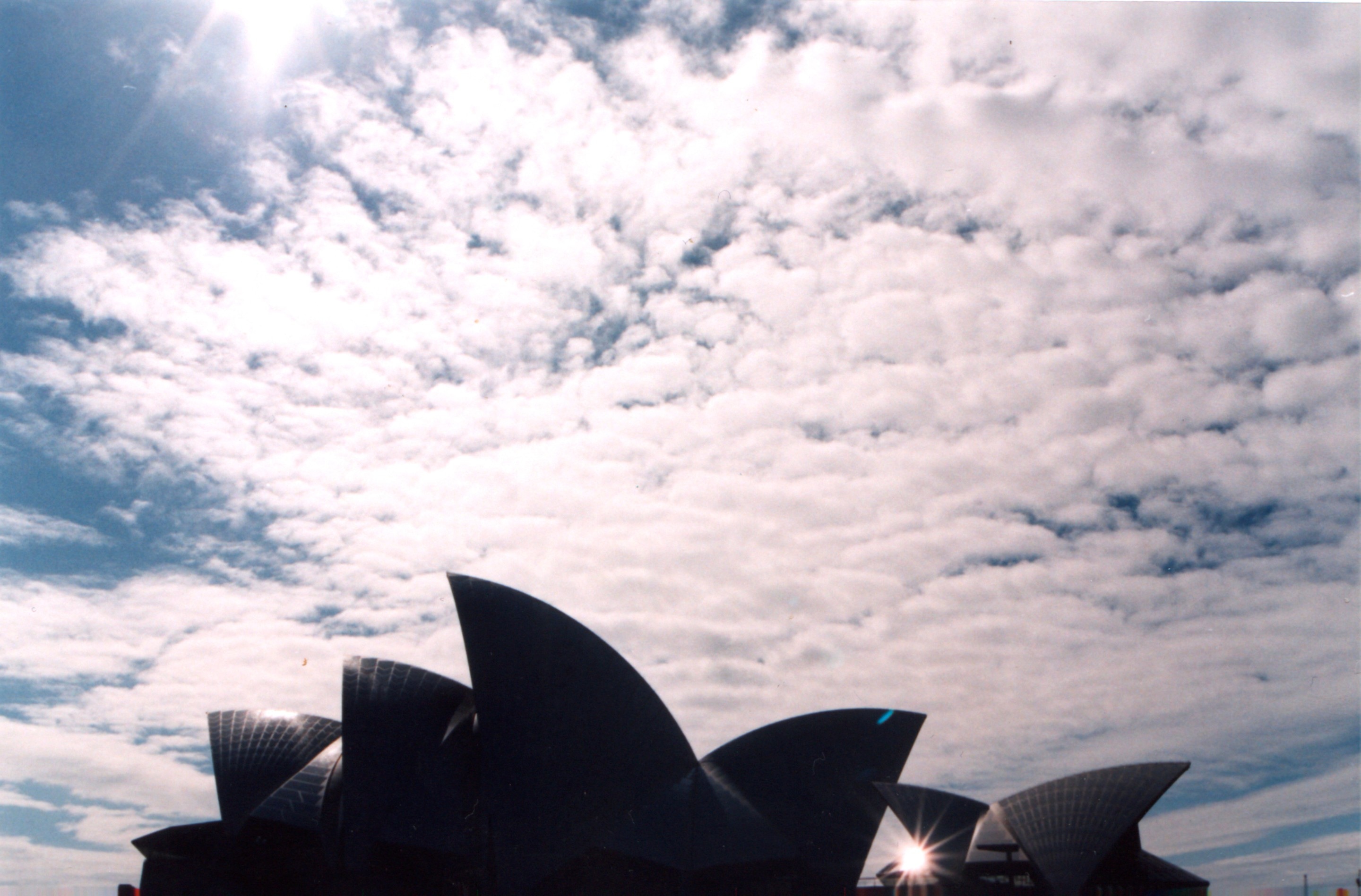 35mm film photograph Sydney Opera House, with clouds above and starburst sun peeping through one of the building's arcs — Sydney, Australia