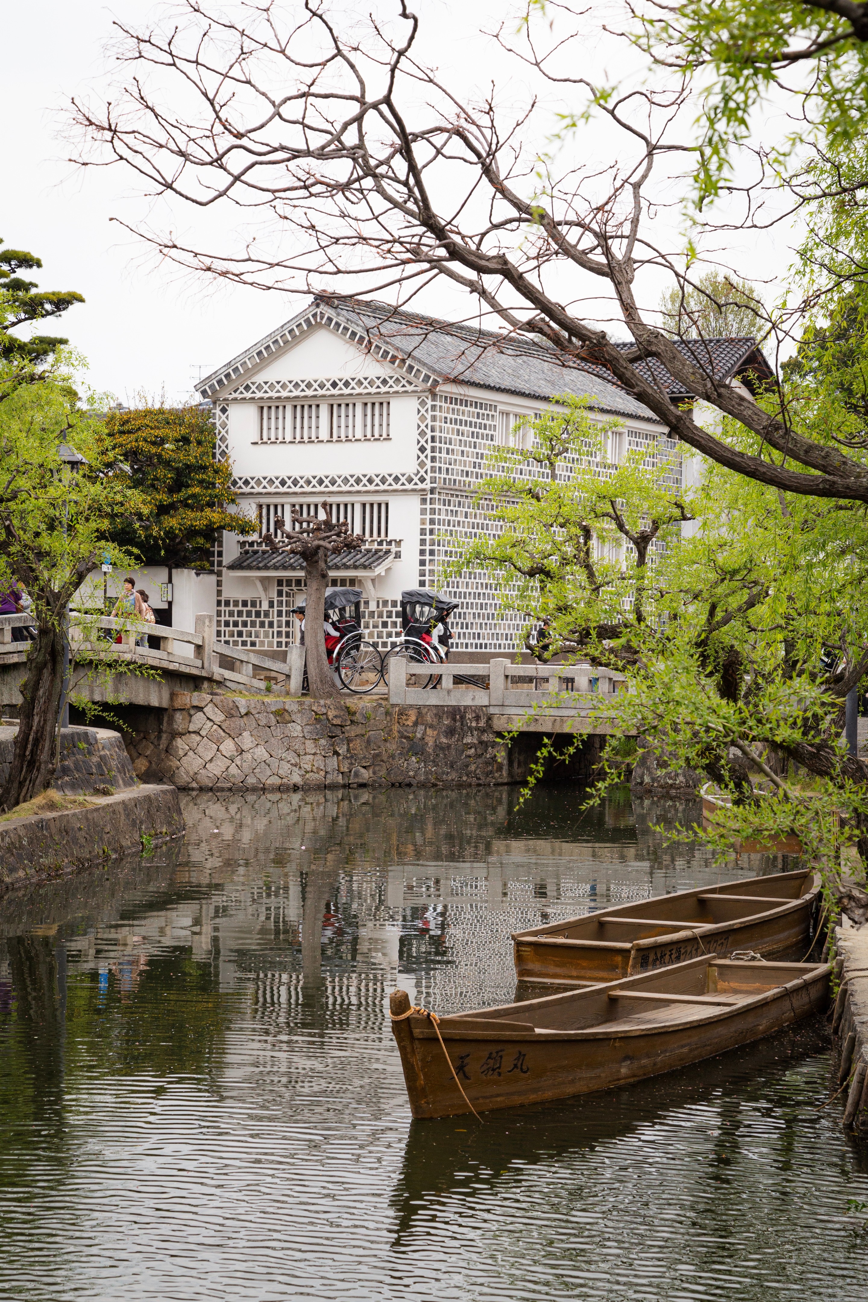 Two wooden rowing boats moored to the side of a stone lined canal with stone pedestrian bridges and two rickshaws in the background — Kurashiki Town, Okayama Prefecture, Japan