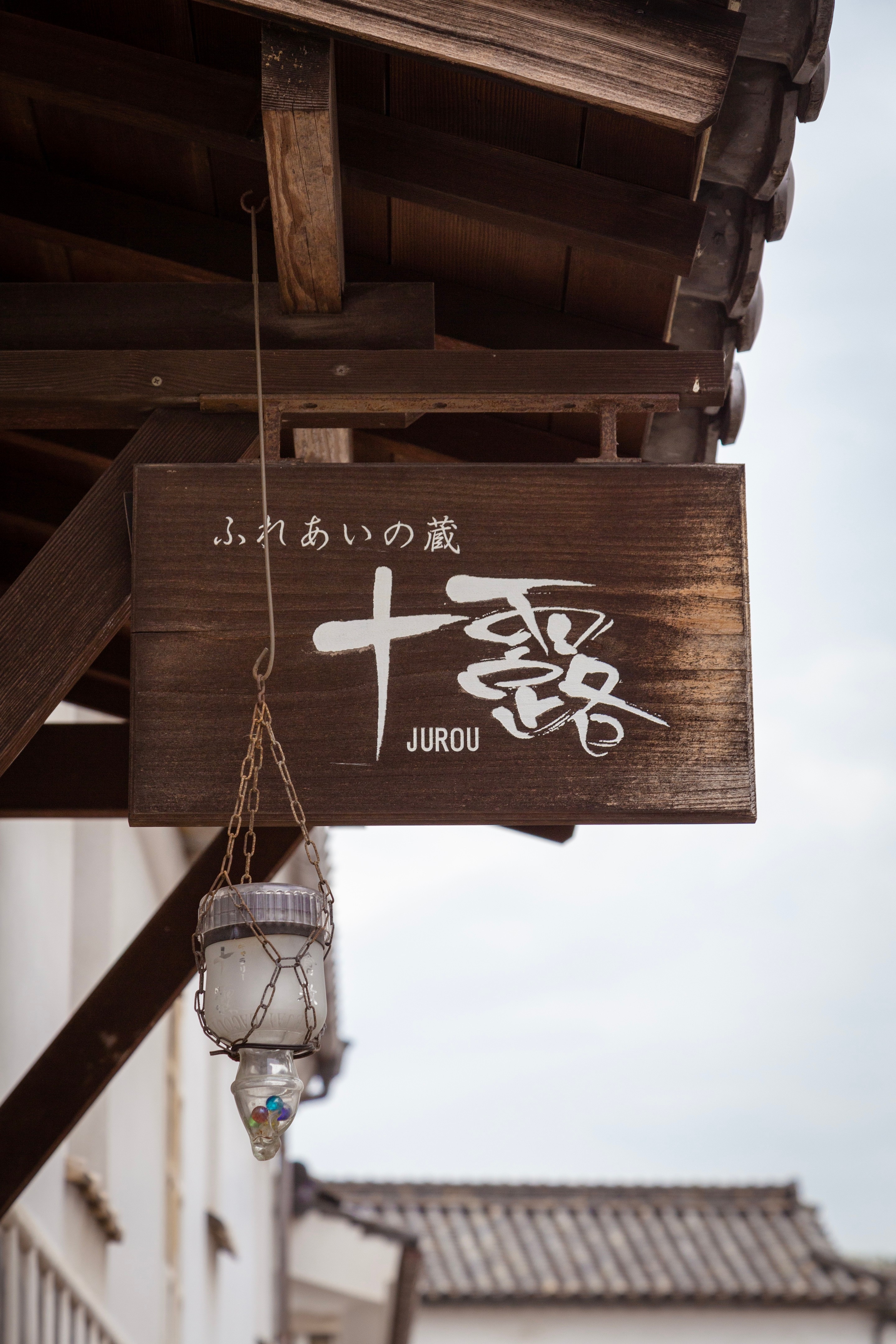 A large wooden shop sigh hangs down from a roof, and attached to it is a glass container holding a few colours spherical glass beads — Kurashiki Town, Okayama Prefecture, Japan