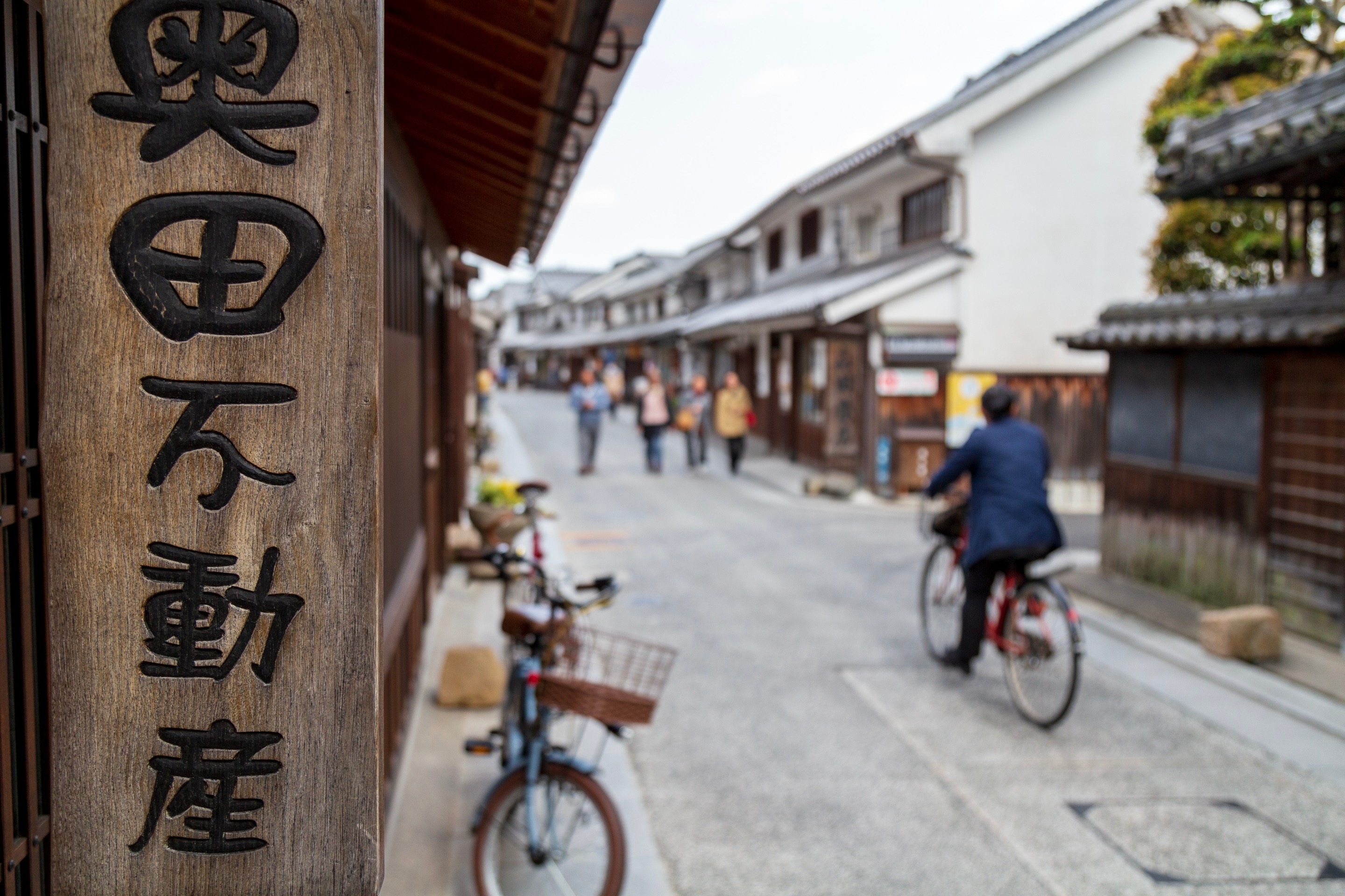 In the foreground a vertical shop sign written in kanji is focused, with the blurred background consisting of a cyclist and people walking along a shopping street in — Kurashiki Town, Okayama Prefecture, Japan