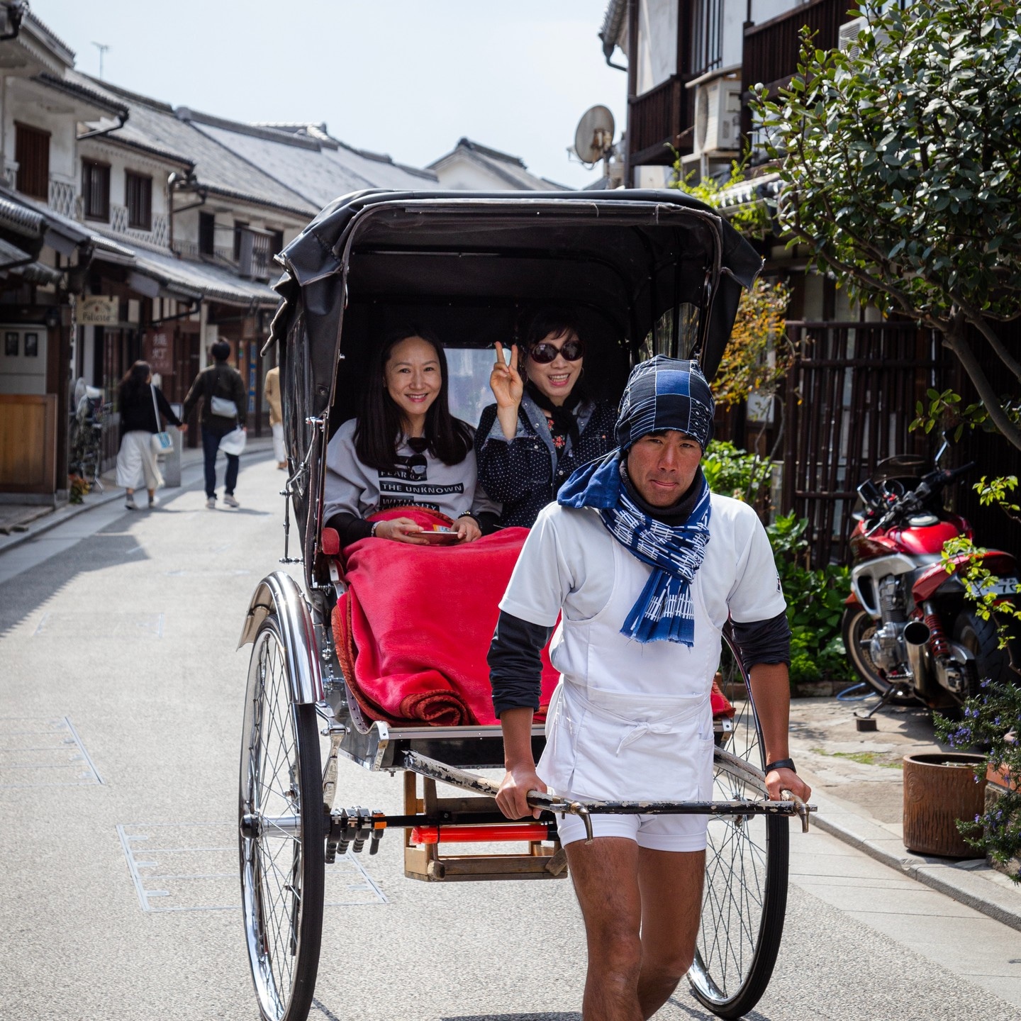 A couple of tourists waving and smiling whilst being taxied in a jinkikisha; rickshaw — along a shopping street in Kurashiki Town, Okayama Prefecture, Japan
