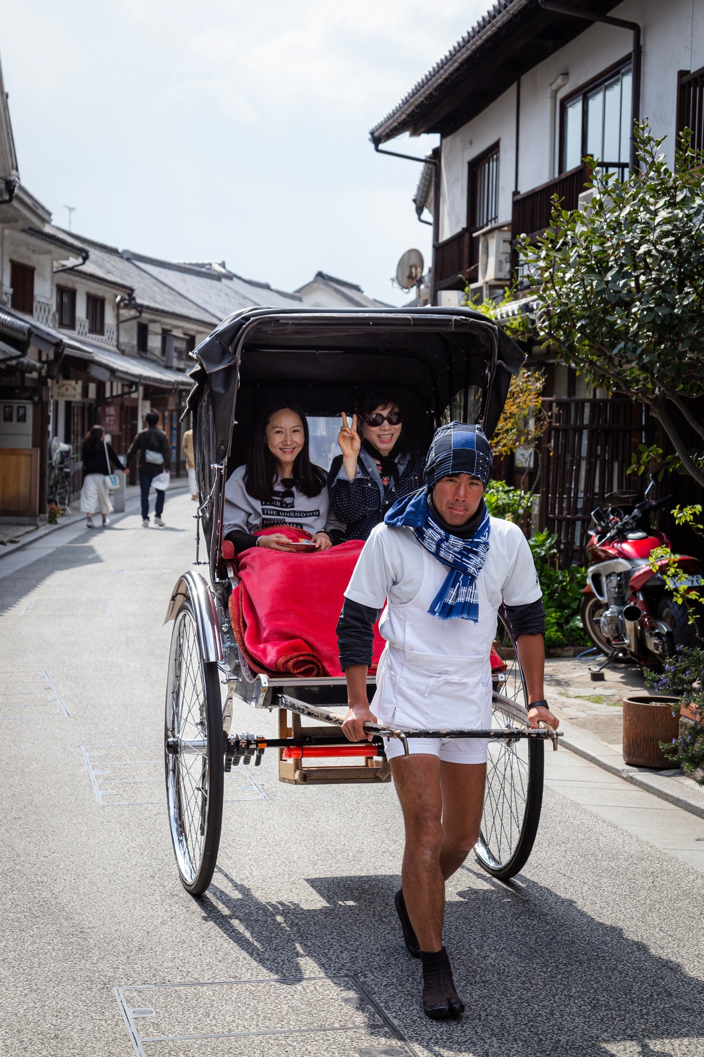 A couple of tourists waving and smiling whilst being taxied in a jinkikisha; rickshaw — along a shopping street in Kurashiki Town, Okayama Prefecture, Japan
