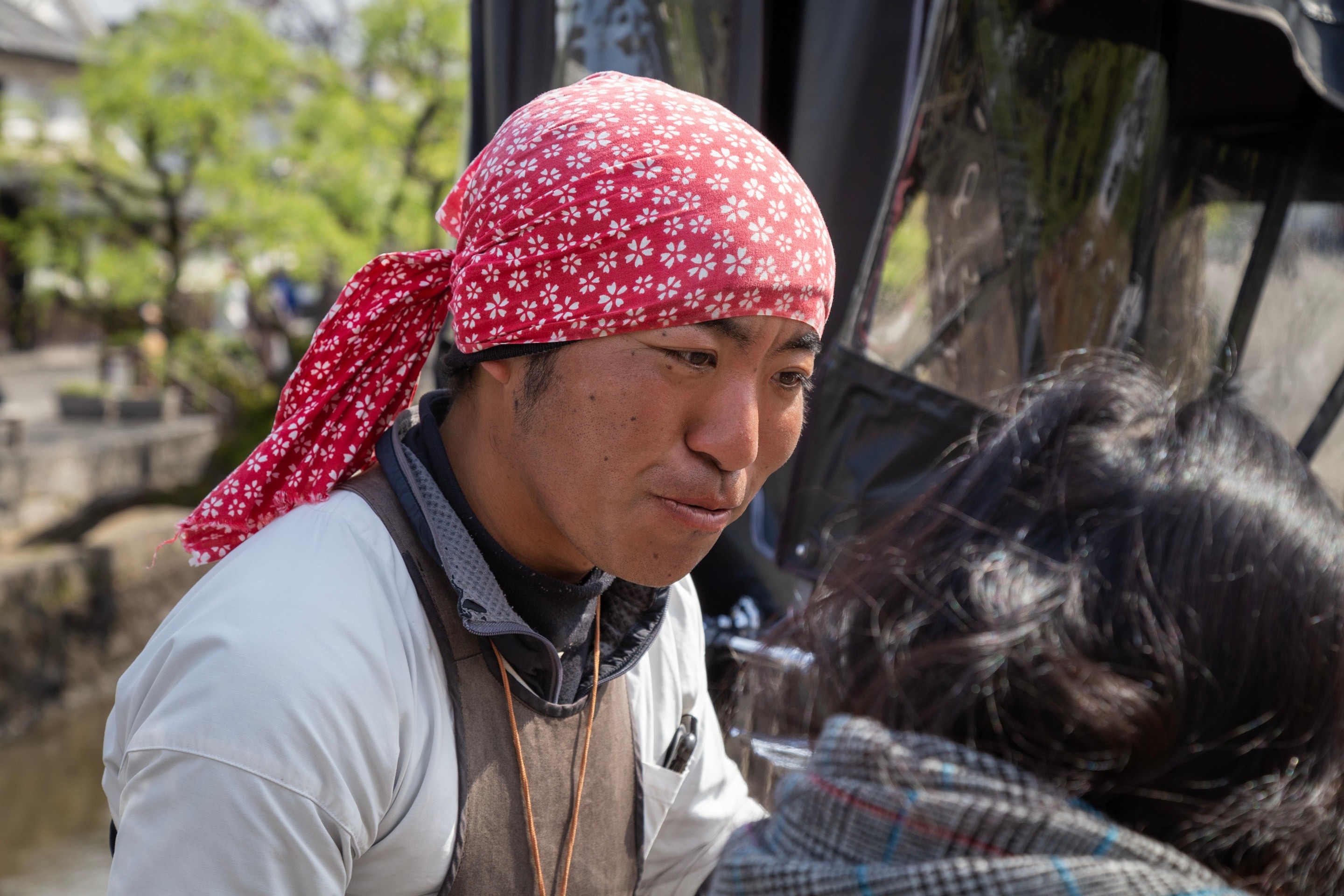 Jinrikisha, or rickshaw, driver wearing a red headscarf with white sakura patterns, talking to an interested tourist with his rickshaw in the background — Kurashiki Town, Okayama Prefecture, Japan