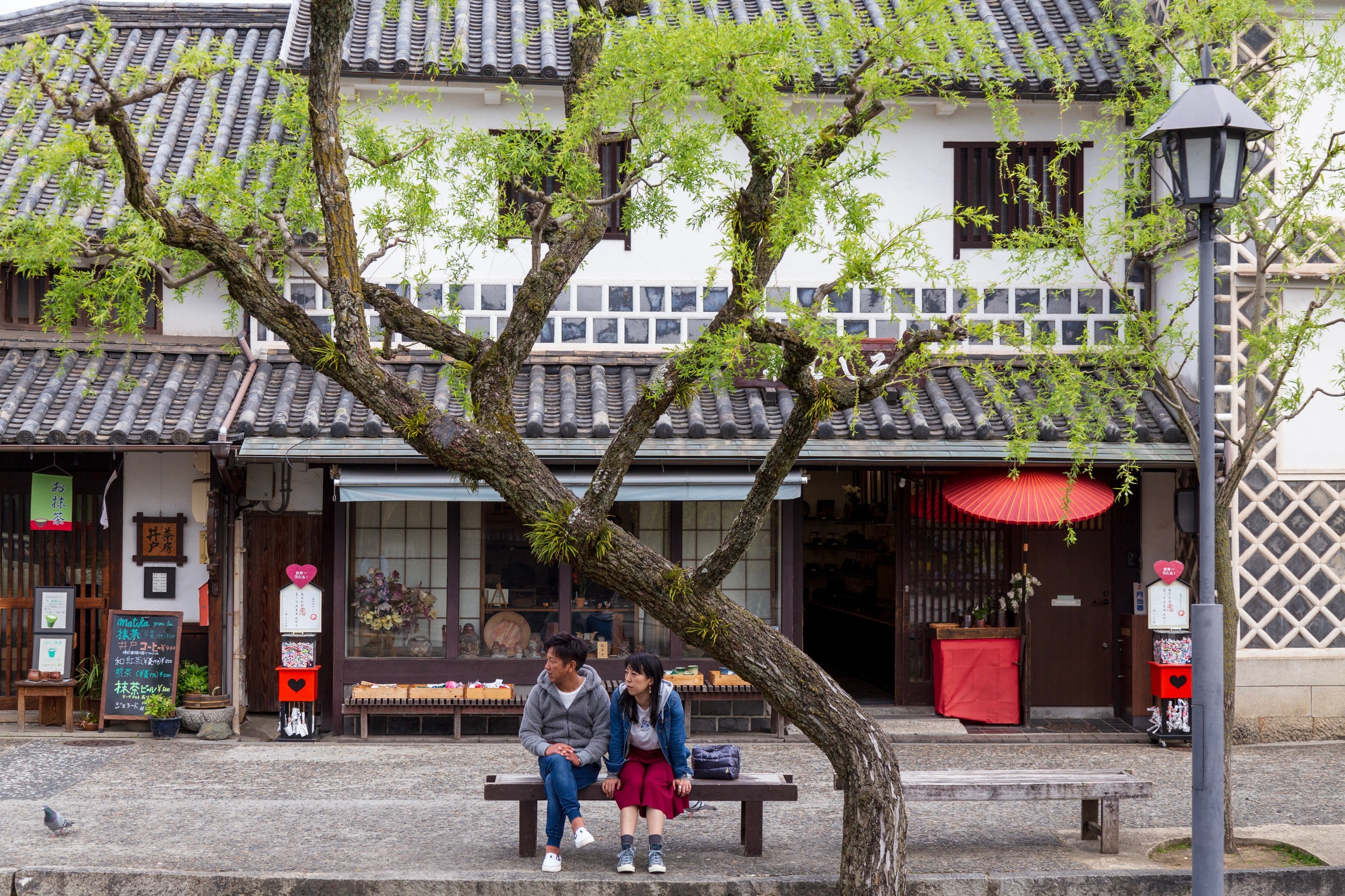 A couple siting on a wooden bench watching the world go by, they are surrounded by willow trees that line a stone canal  — Kurashiki Town, Okayama Prefecture, Japan
