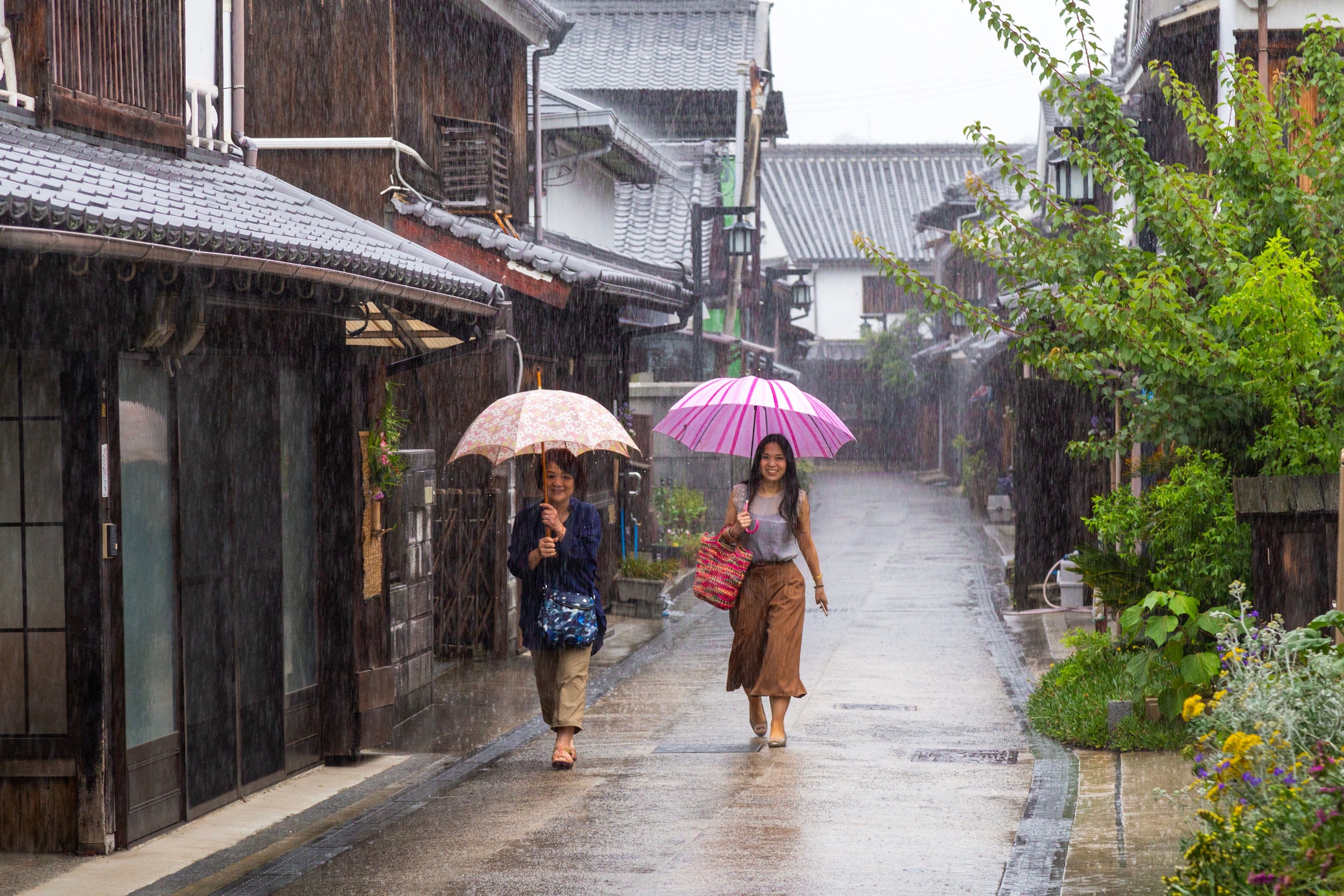 Two woman waking down an old traditional street in drizzly conditions holding opened umbrellas — Mitarai village, Osaki Shimojima Island, Geiyo Region.