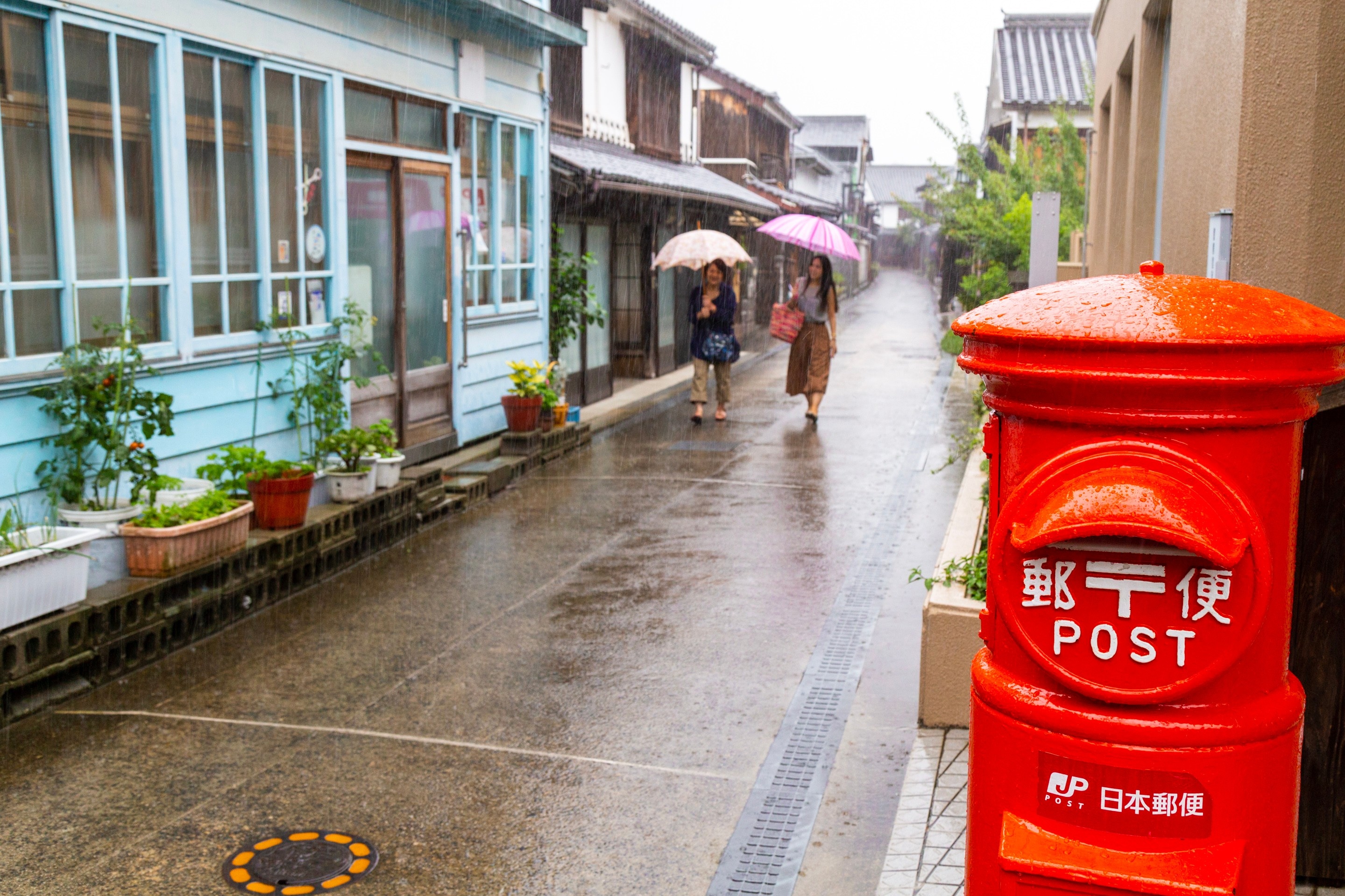 Two woman waking down an old traditional street in drizzly conditions holding opened umbrellas in the background, with a bright red postbox in the foreground — Mitarai village, Osaki Shimojima Island, Geiyo Region.