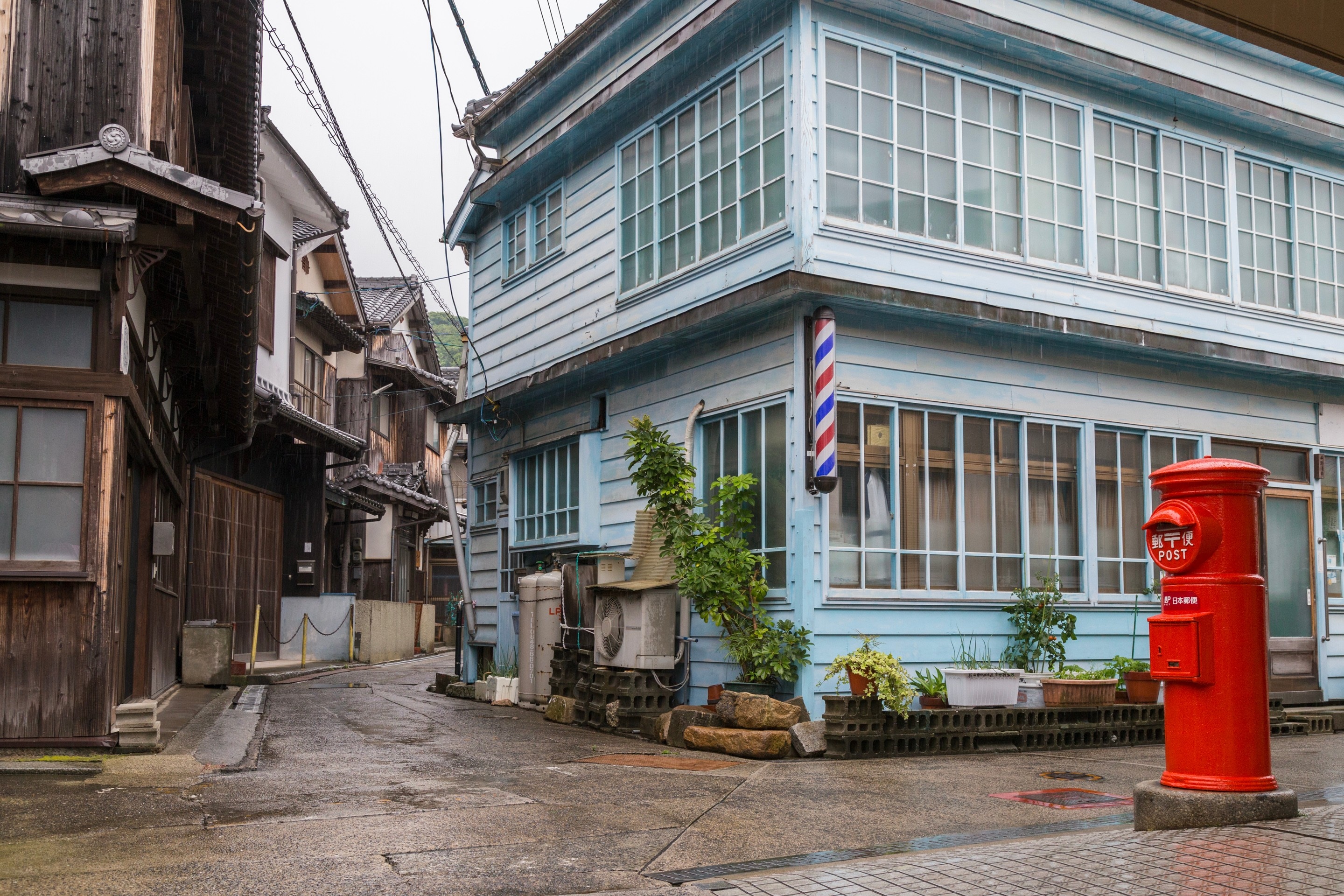 A large painted blue storehouse building flanks a narrow alley filled with traditional wooden residential town houses; in the foreground to the right is a bright red postbox — Mitarai village, Osaki Shimojima Island, Geiyo Region.