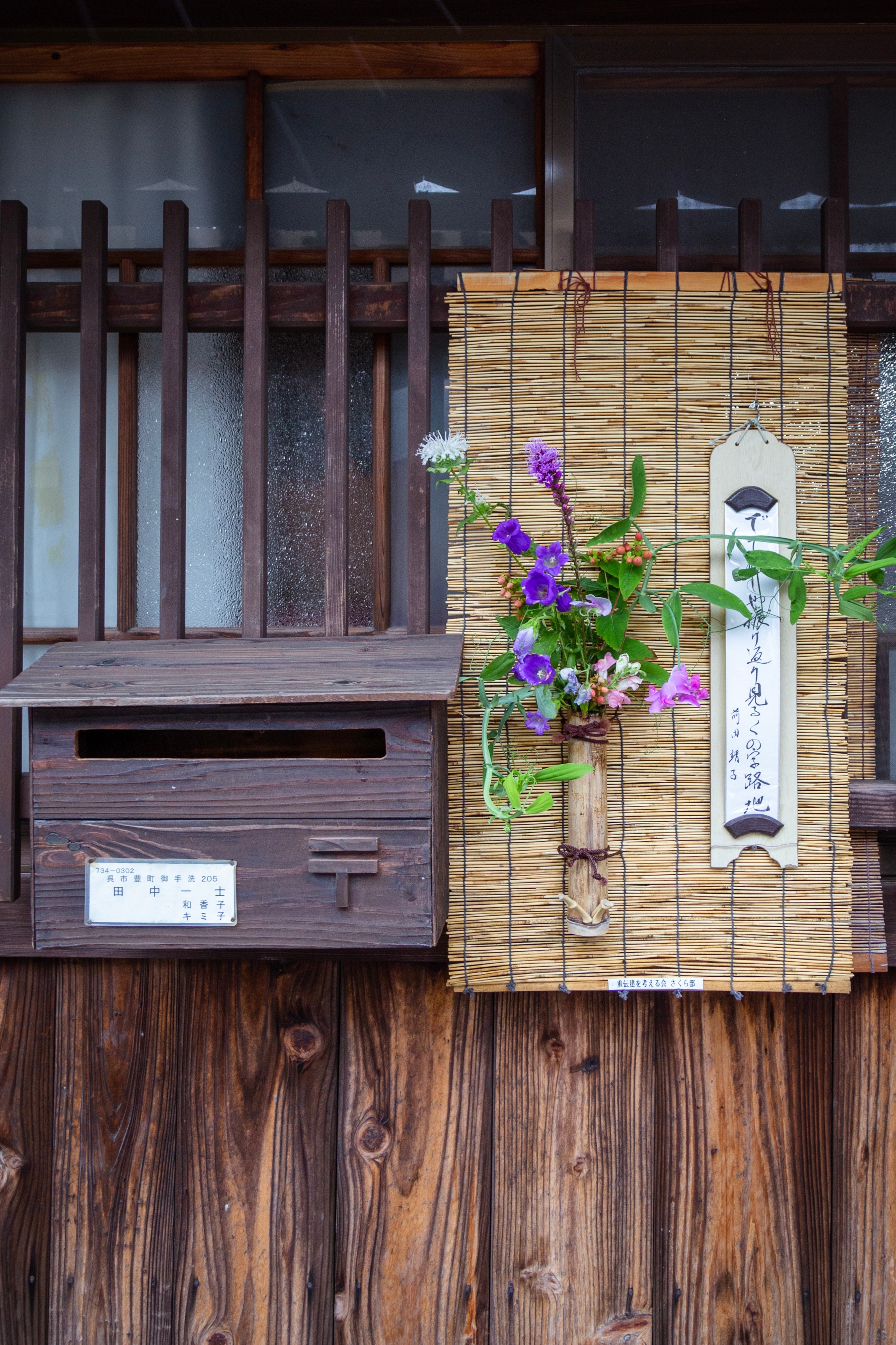 Locals adorn their entrances with bouquets of flowers and poems to welcome visitors to the island and village. Flowers are in a bamboo vase attached to a small bamboo blind, next to which is a postbox and a short poem — Mitarai, Geiyo Region, Japan