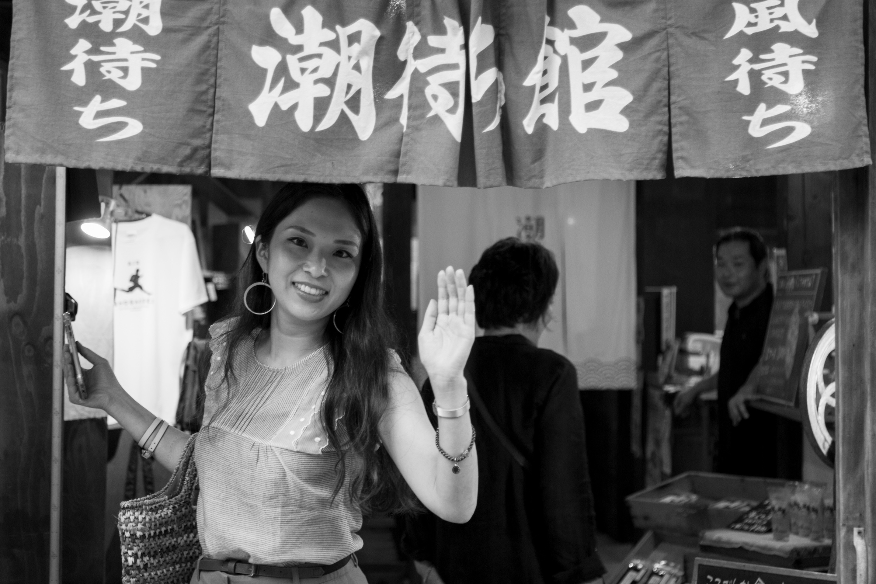 A young Japanese woman waves at the camera from a shop entrance, above her are norem; traditional entrance curtains with sinographs printed on them — Mitarai village, Osaki Shimojima Island, Geiyo Region.