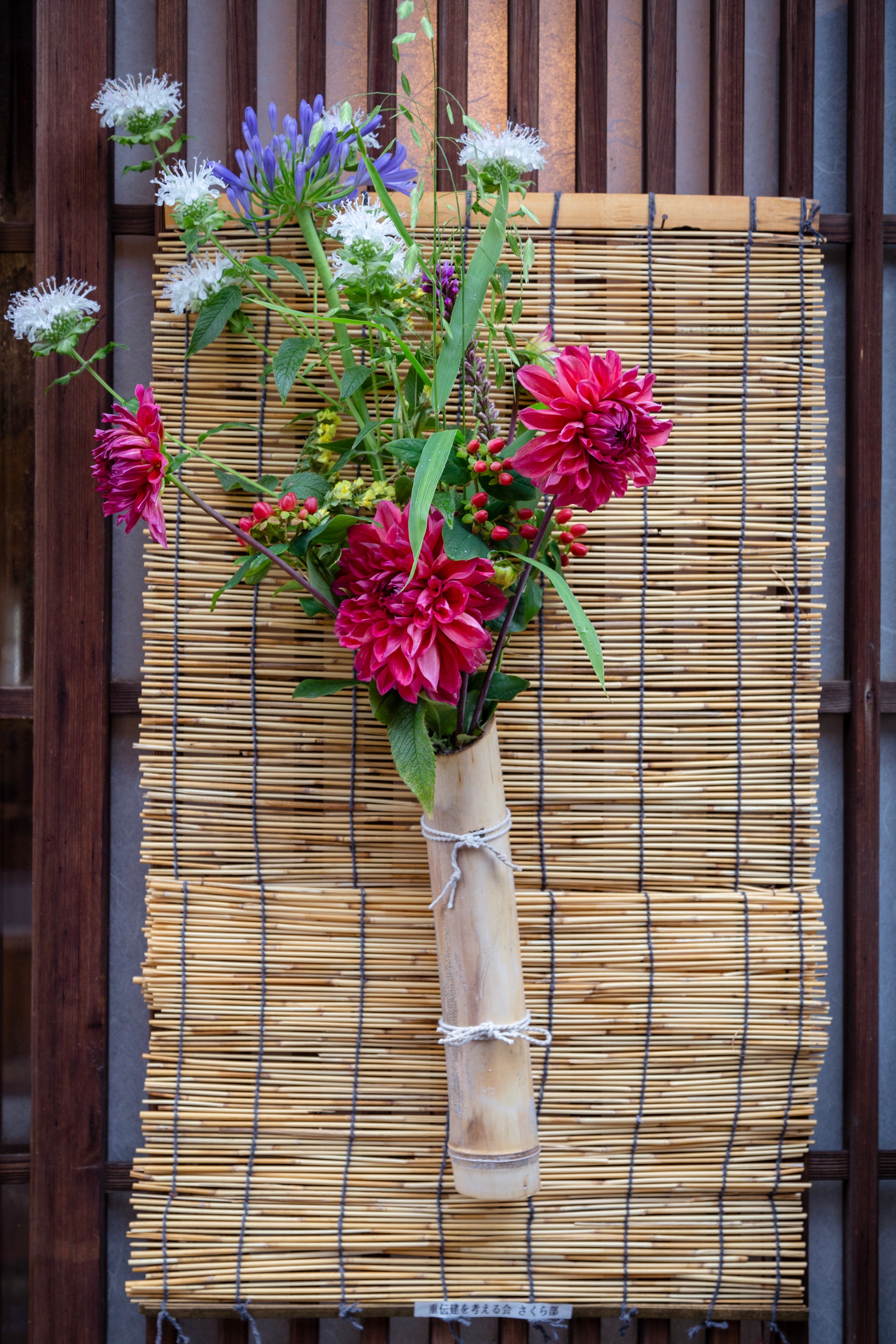Locals adorn their entrances with bouquets of flowers and poems to welcome visitors to the island and village. Colourful flowers are in a bamboo vase attached to a small bamboo blind — Mitarai, Geiyo Region, Japan