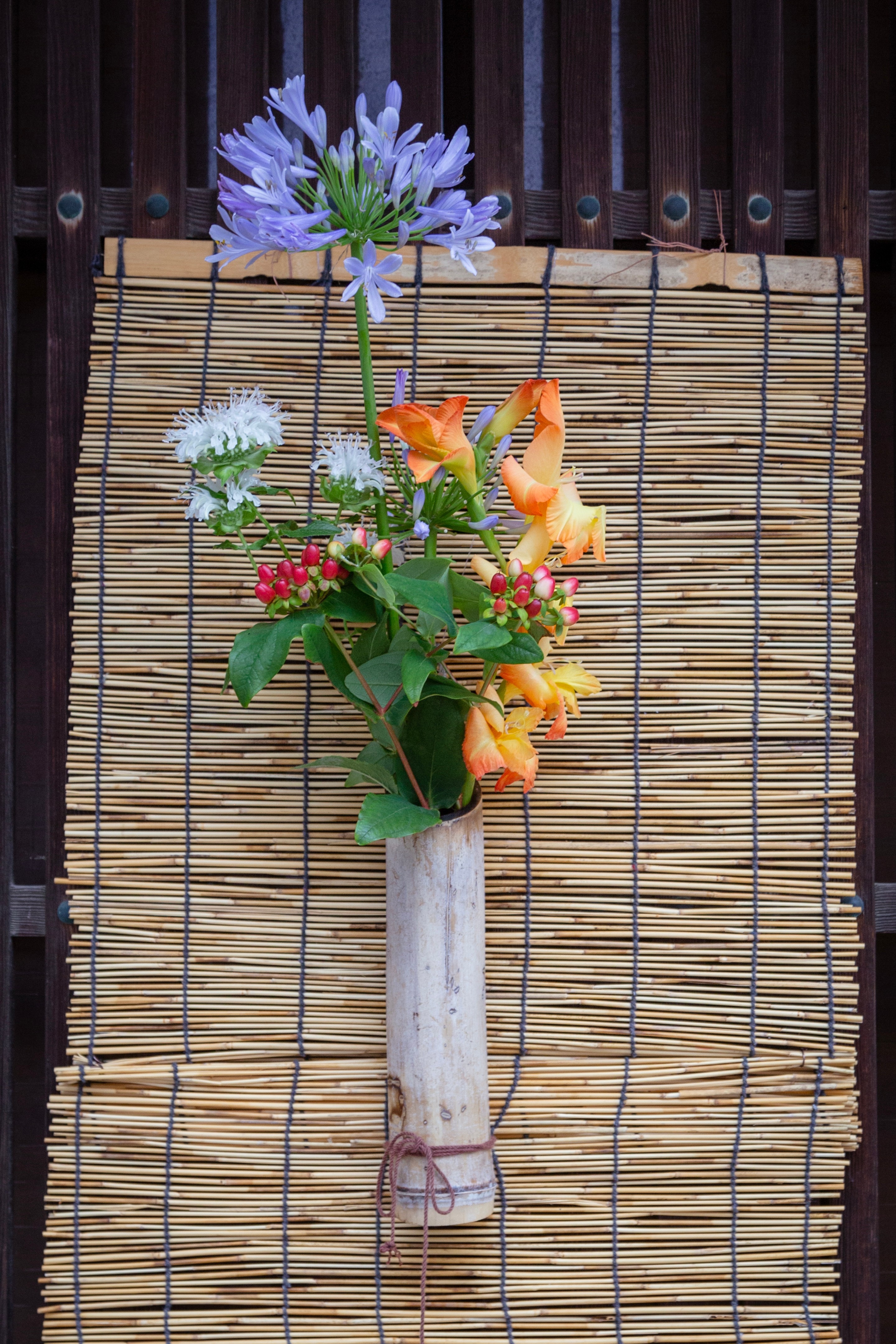 Locals adorn their entrances with bouquets of flowers and poems to welcome visitors to the island and village. Colourful flowers are in a bamboo vase attached to a small bamboo blind — Mitarai, Geiyo Region, Japan