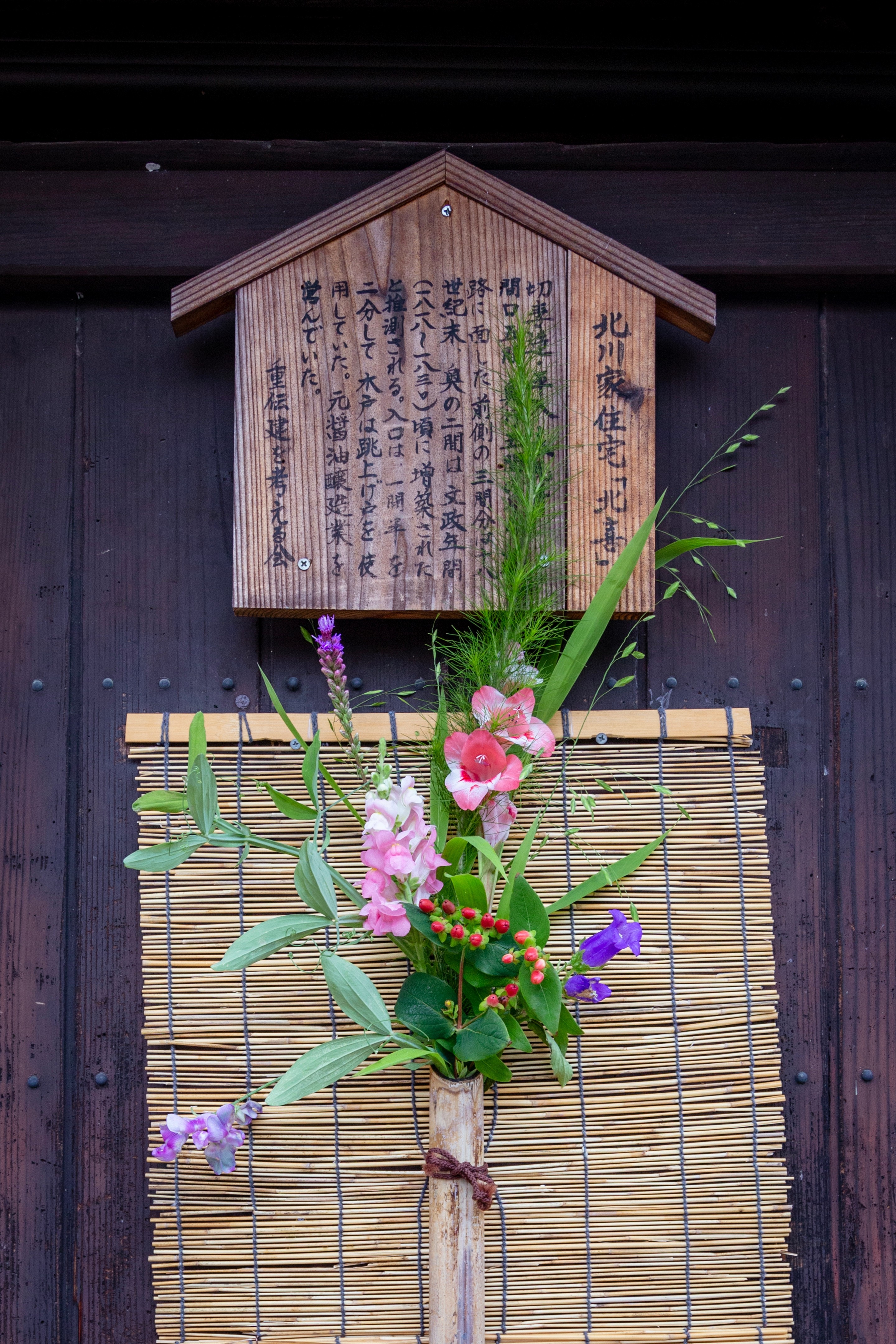 Locals adorn their entrances with bouquets of flowers and poems to welcome visitors to the island and village. Flowers are in a bamboo vase attached to a small bamboo blind, above which is a long poem carved into a wooden panel — Mitarai, Geiyo Region, Japan