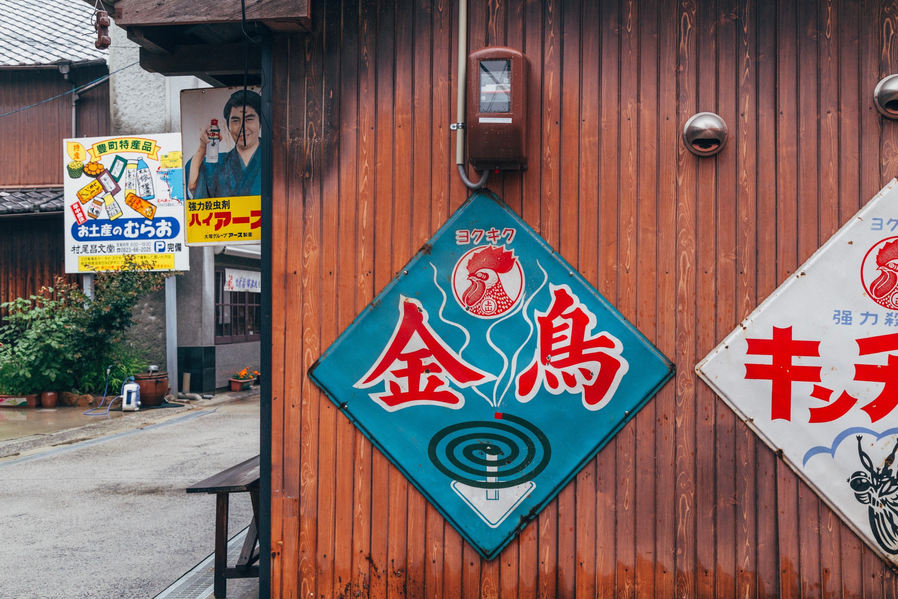 Large old metal signs nailed to the side of a wooded shop advertising insect repellant coils and insect killer spray. A traditional street can be seen to the left side — Mitarai village, Osaki Shimojima Island, Geiyo Region.