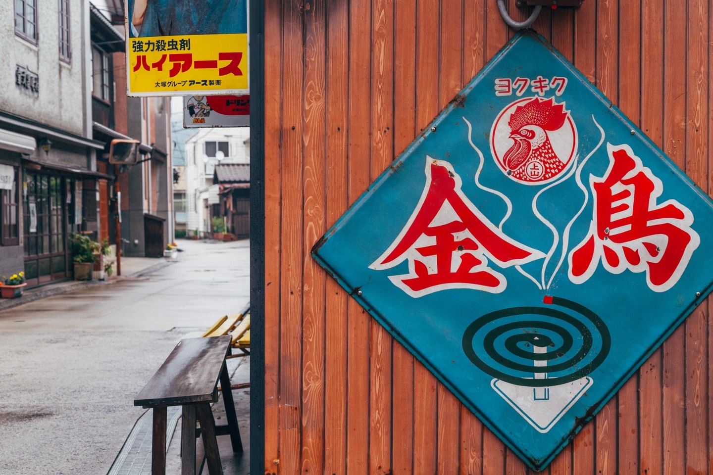 Large old metal signs nailed to the side of a wooded shop advertising insect repellant coils and insect killer spray. A traditional street can be seen to the left side — Mitarai village, Osaki Shimojima Island, Geiyo Region, Japan