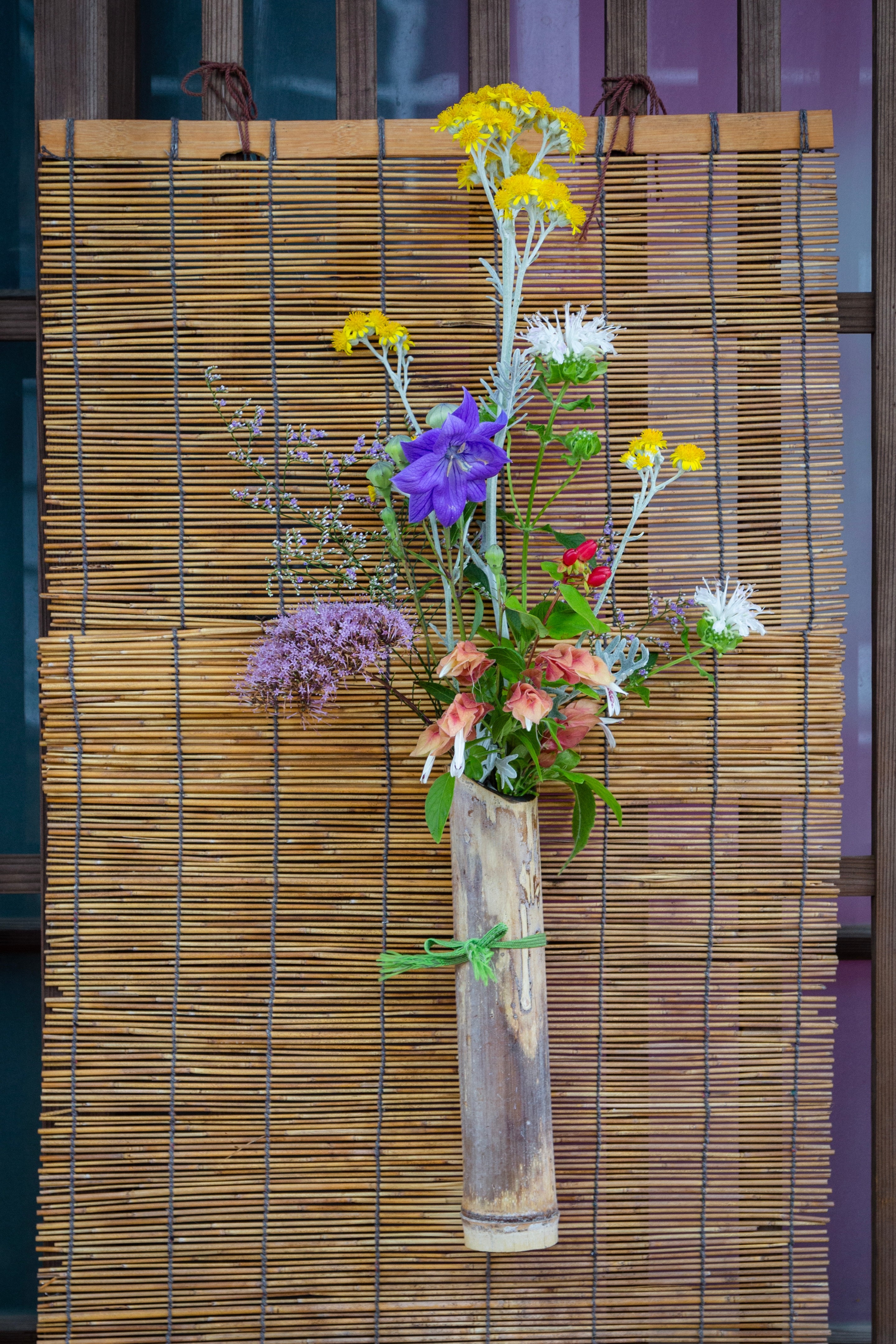 Locals adorn their entrances with bouquets of flowers and poems to welcome visitors to the island and village. Colourful flowers are in a bamboo vase attached to a small bamboo blind — Mitarai, Geiyo Region, Japan