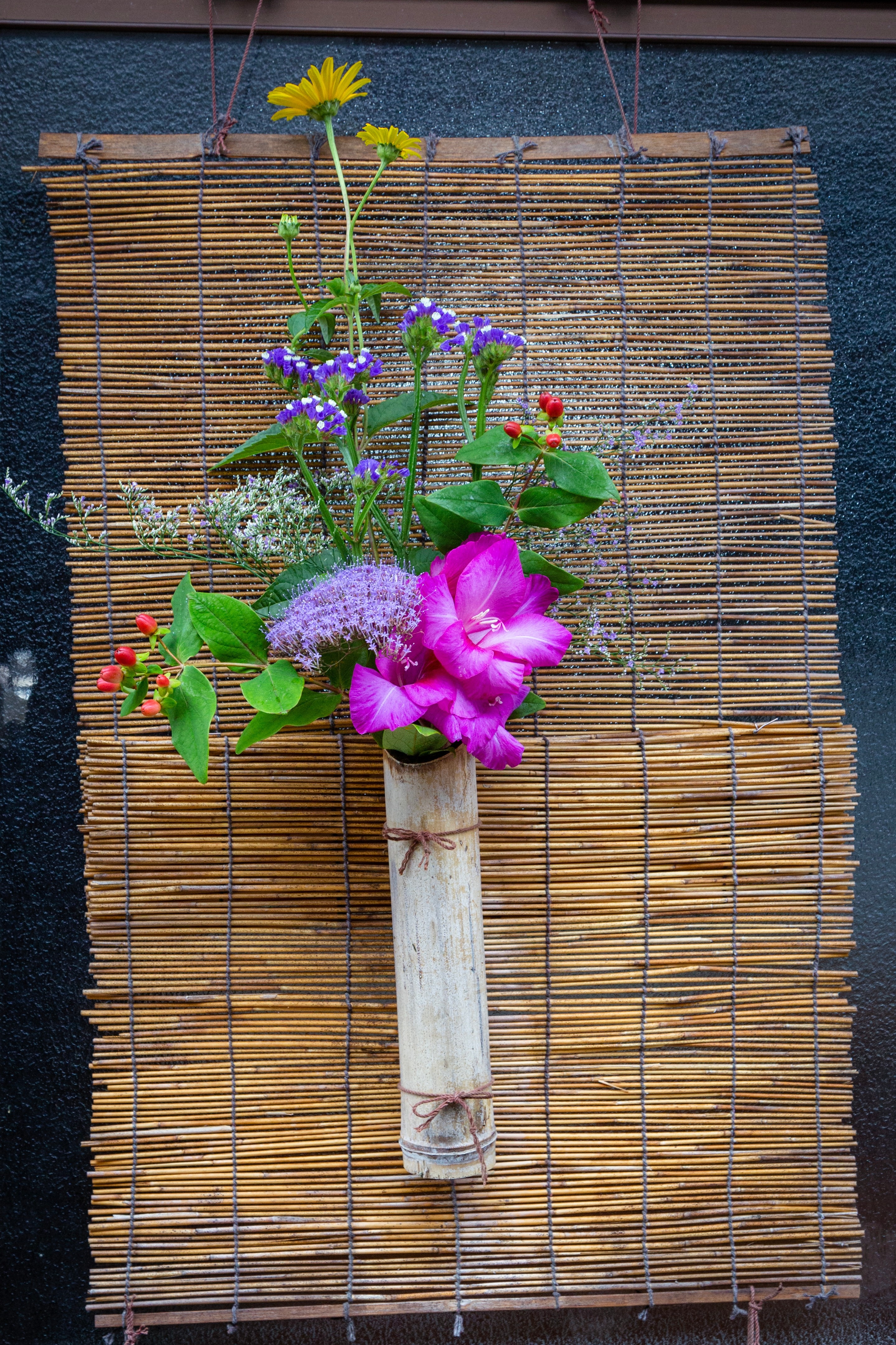 Locals adorn their entrances with bouquets of flowers and poems to welcome visitors to the island and village. Colourful flowers are in a bamboo vase attached to a small bamboo blind — Mitarai, Geiyo Region, Japan