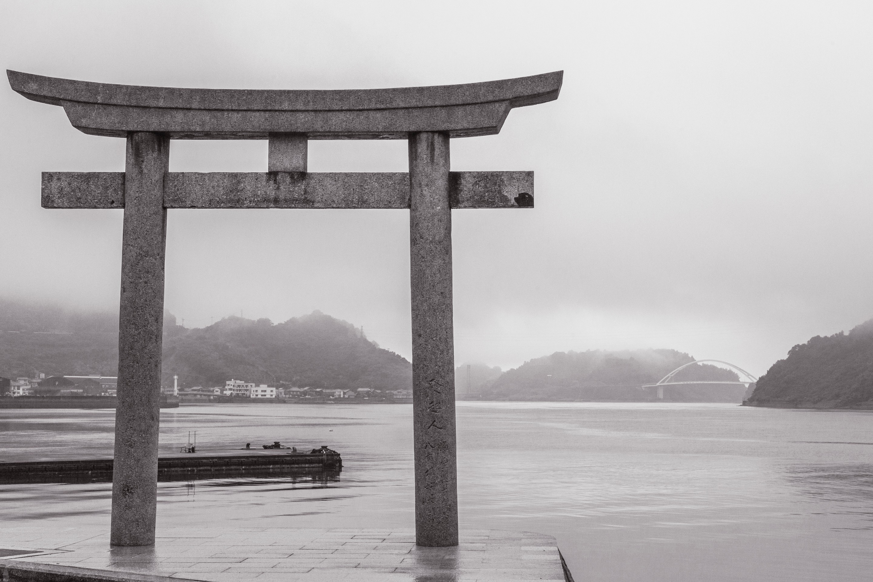 A stone Shinto Torii; Japanese spiritual gate with the ocean, islands and bridge spanning two islands seen the background in black and white — Mitarai village, Osaki Shimojima Island, Geiyo Region.