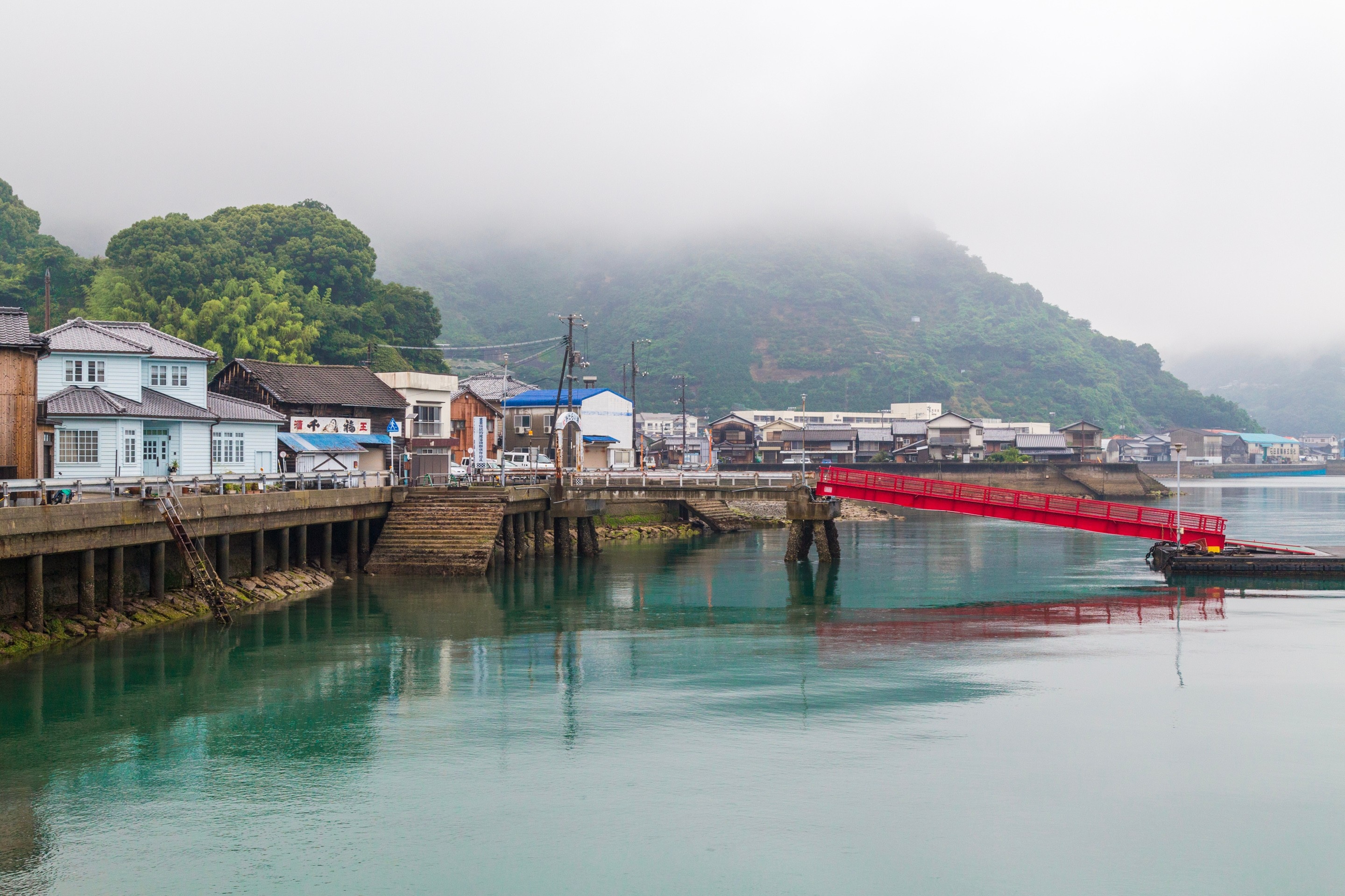 A coastal high street and a bright red gangway lowered onto a pontoon surrounded by aquamarine sea water in drizzly conditions — Mitarai village, Osaki Shimojima Island, Geiyo Region.