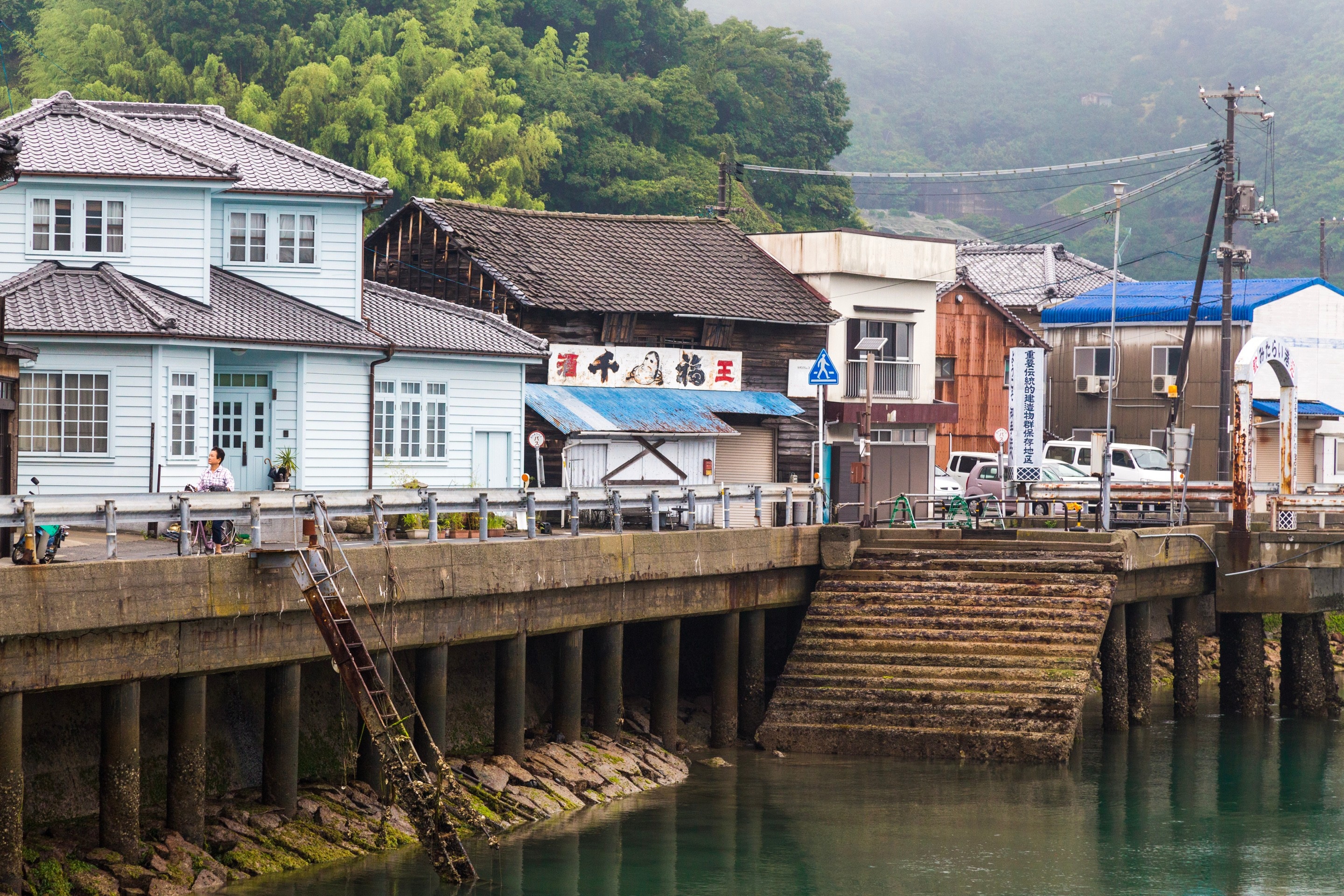 A coastal high street with shops and warehouses with signage, a sole person rides a pink bicycle in drizzly conditions — Mitarai village, Osaki Shimojima Island, Geiyo Region.