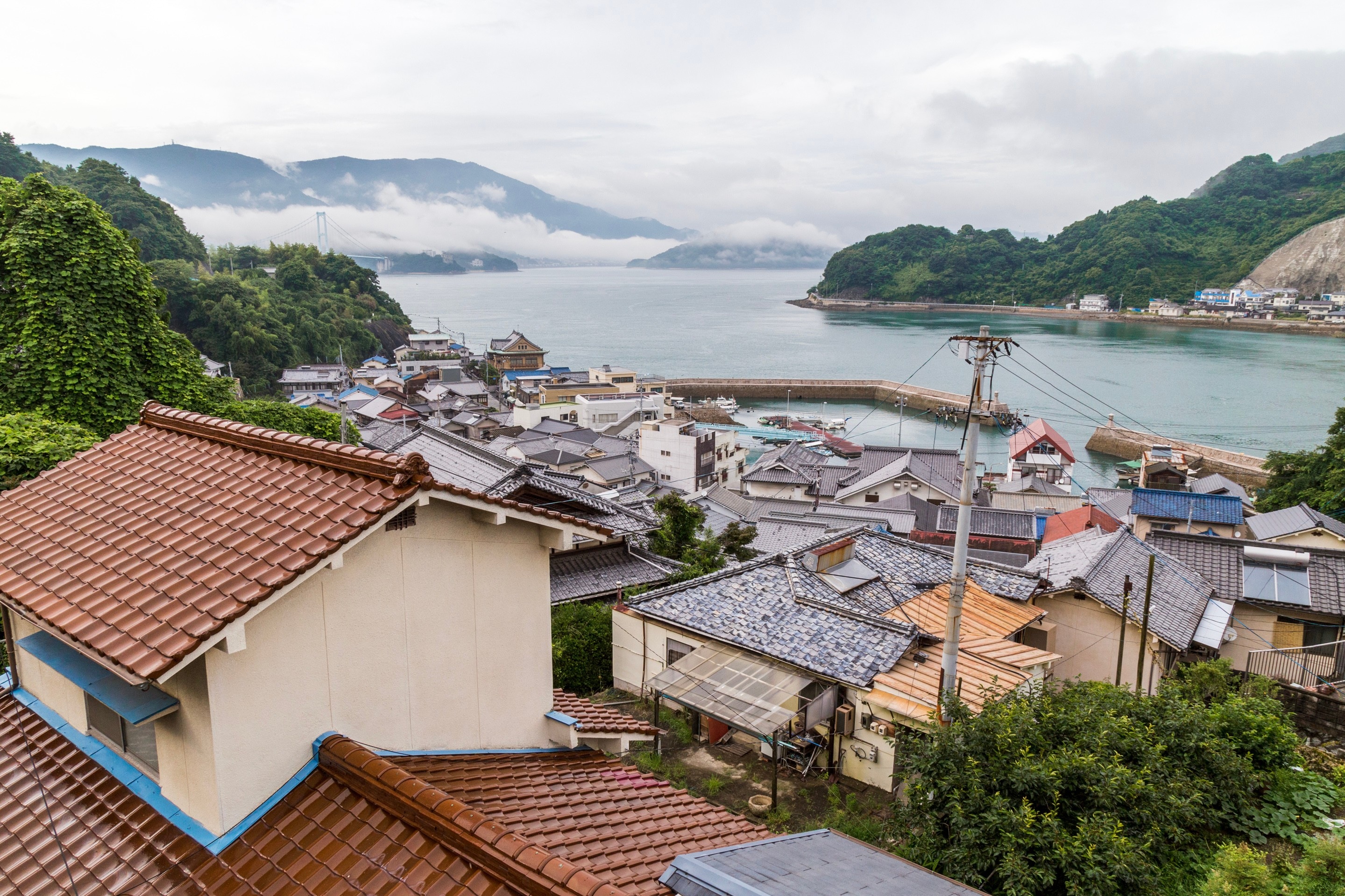 Looking down over tiled rooftops of residential homes, with cloud obscured islands, aquamarine ocean and a blue suspension bridge in the background — Mitarai village, Osaki Shimojima Island, Geiyo Region.