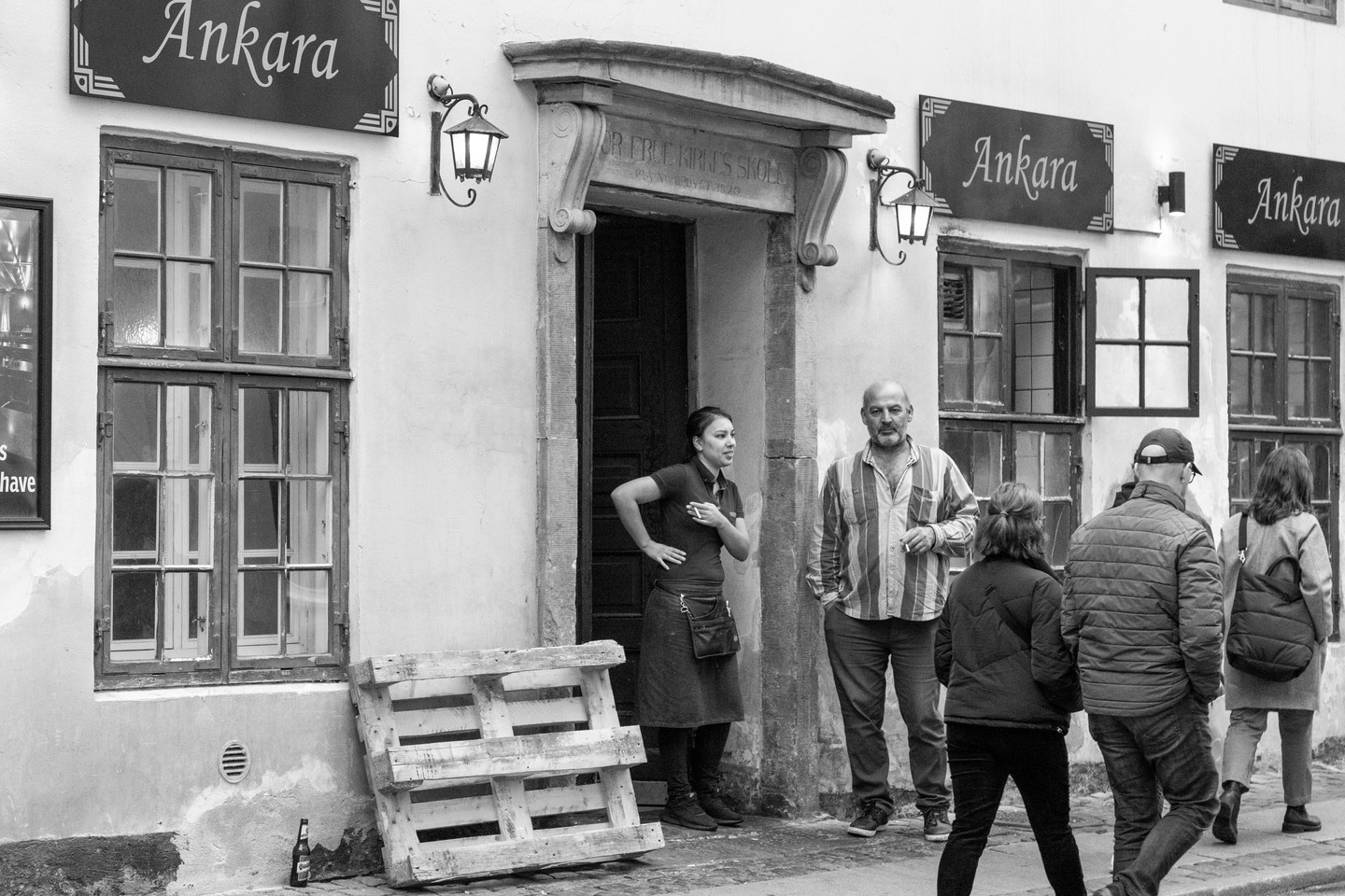 Black and white photo of a two restaurant workers enjoying a smoking break, standing talking within the entrance to the establishment. The man on the right is staring directly into the lens. Copenhagen, Denmark