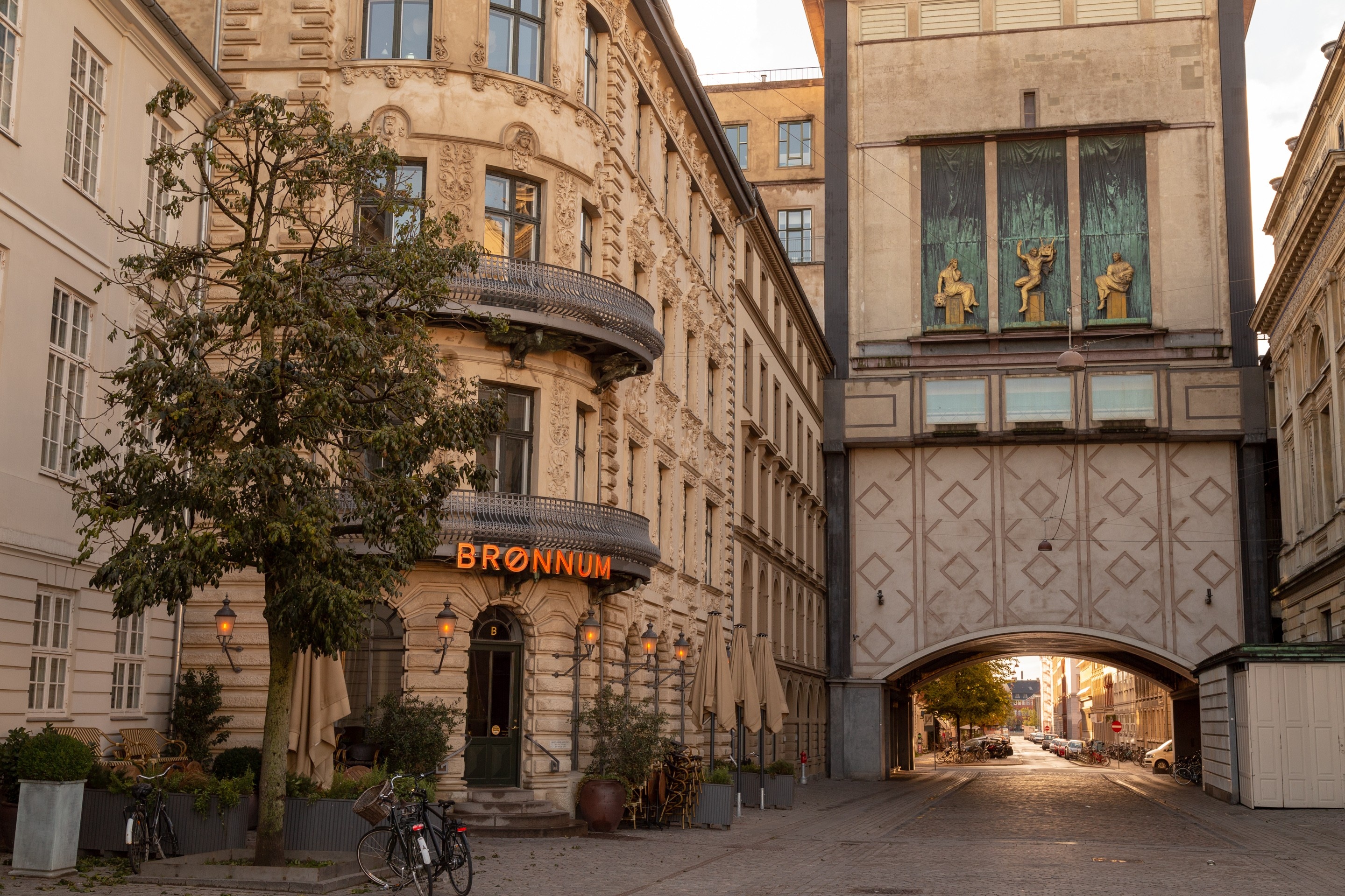 Tranquil evening city scene during golden hour. To the left an elegant orange neon signs glows invitingly above an entrance to a restaurant. To the right, the eye is led under an archway that lead to an illuminated golden hour street. Copenhagen, Denmark.