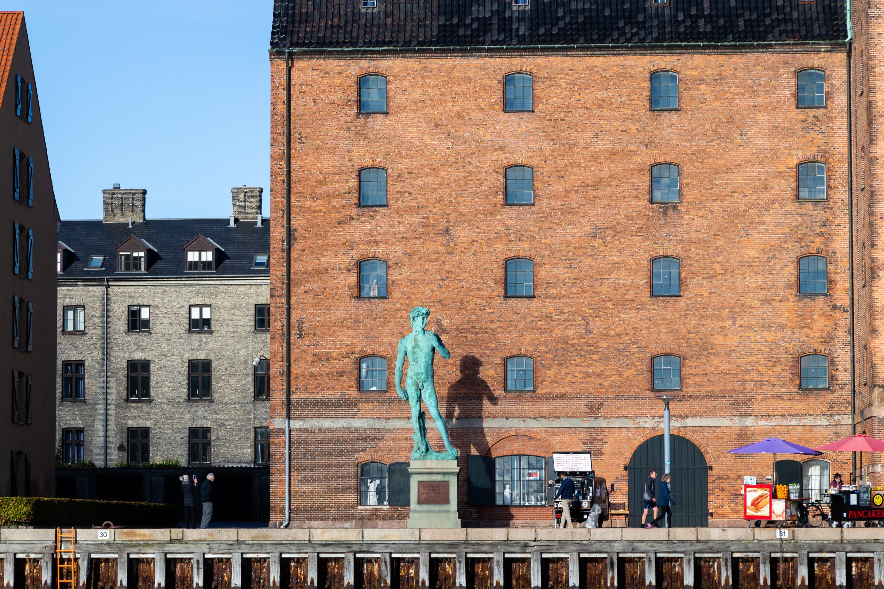 A copy of the original David sculpture by Michelangelo. This one in bronze, which has aged beautifully to vibrant hues of green and blue. The statue sits along the waterfront area of Copenhagen Harbour. Immediately behind the statue is a grand brick warehouse, converted to offices. Denmark.