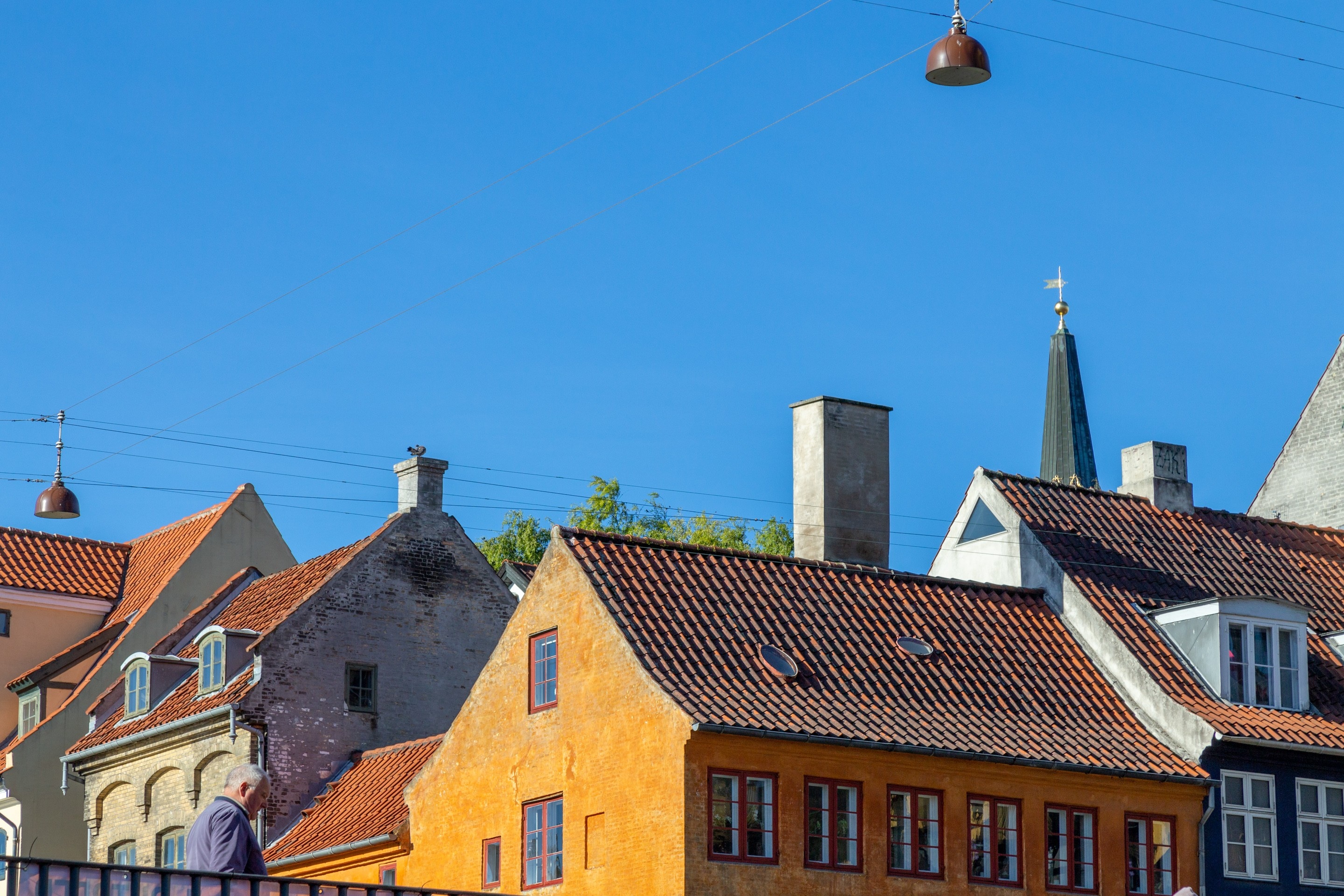 Layers of colourful earthy and orange townhouses fill the lower half of the scene, with a rich blue sky filling up the remainder. A man can be seen to the bottom left crossing a bridge. Copenhagen, Denmark.
