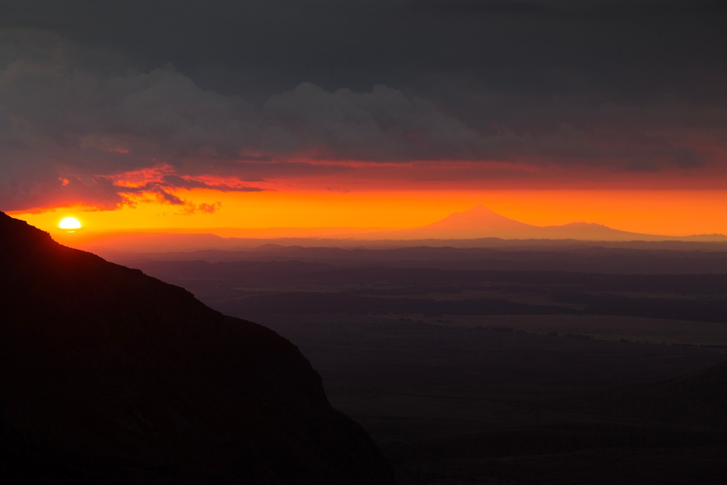 A beautiful dusk scene taken from Mount Ngauruhoe (Mount Doom) looking out across the North Island to Mount Taranaki. A serene band of orange and peach colours from the setting sun illuminate the central scene — Tongariro National Park, New Zealand