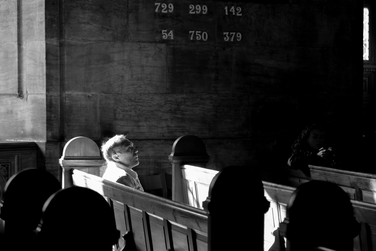 A man is caught in deep contemplation whilst sitting amongst the pews within a church. He is illuminated from behind by a strong shard of light, emphasising his presence within the darker scene around him. Copenhagen, Denmark.