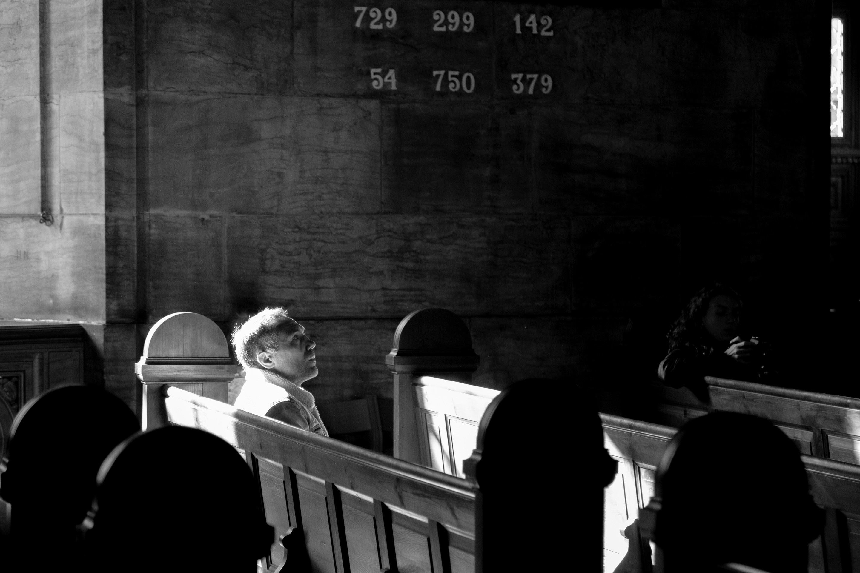 A man is caught in deep contemplation whilst sitting amongst the pews within a church. He is illuminated from behind by a strong shard of light, emphasising his presence within the darker scene around him. Copenhagen, Denmark.