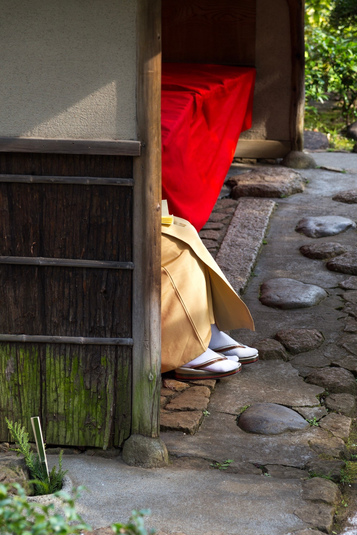 A woman sits and reads whilst sitting on a red velvet blanket inside a bamboo seating area — her face and body obscured by the building's wall, with only her sandals and kimono visible — Nezu Museum, Minato, Tokyo