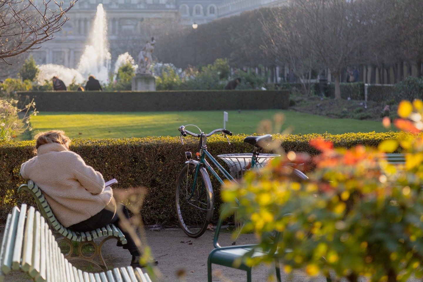 A woman sits on a bench during golden hour, reading a book, with her bicycle resting against another bench. A park and fountain can be seen beyond — Paris, France