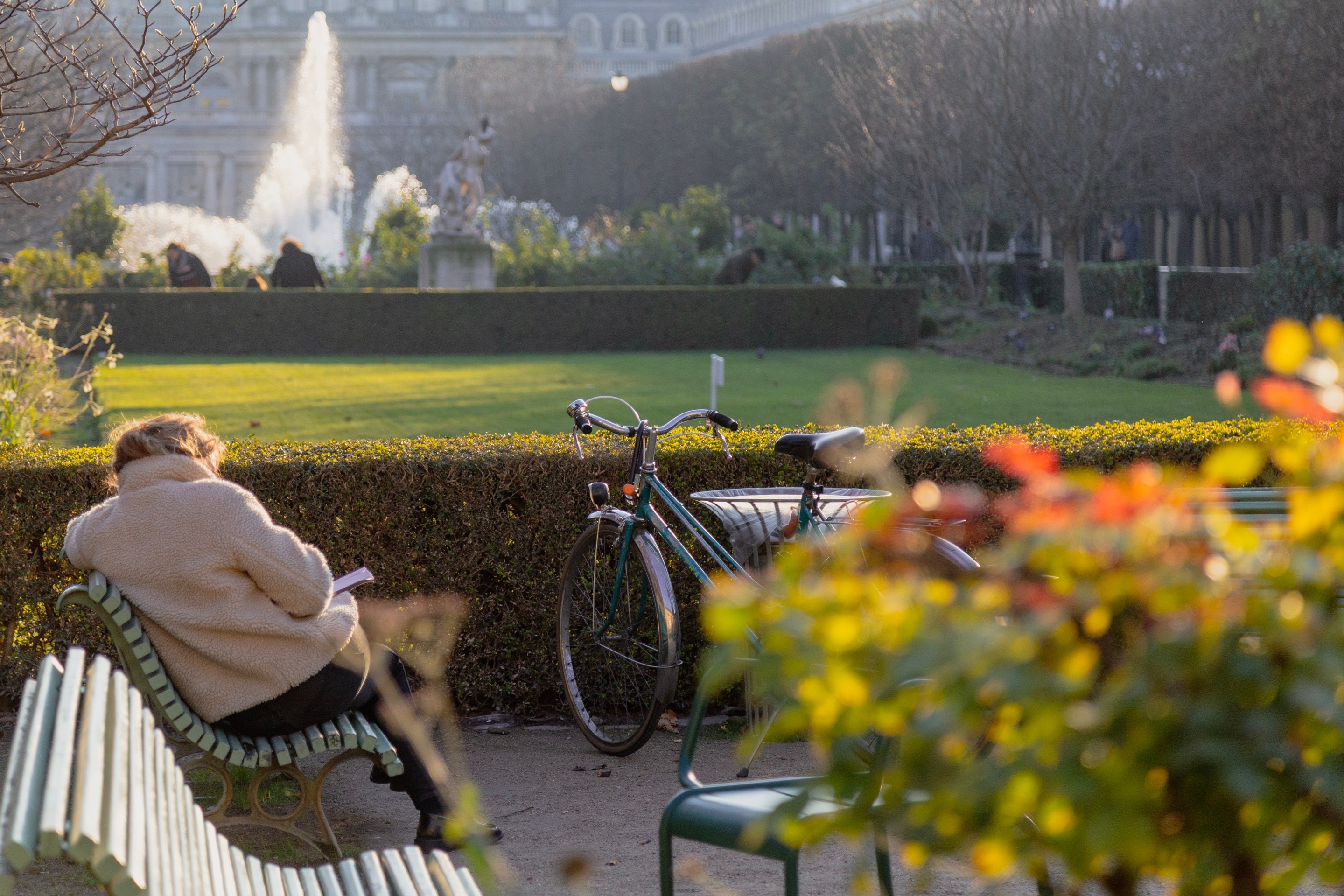 A woman sits on a bench during golden hour, reading a book, with her bicycle resting against another bench. A park and fountain can be seen beyond — Paris, France