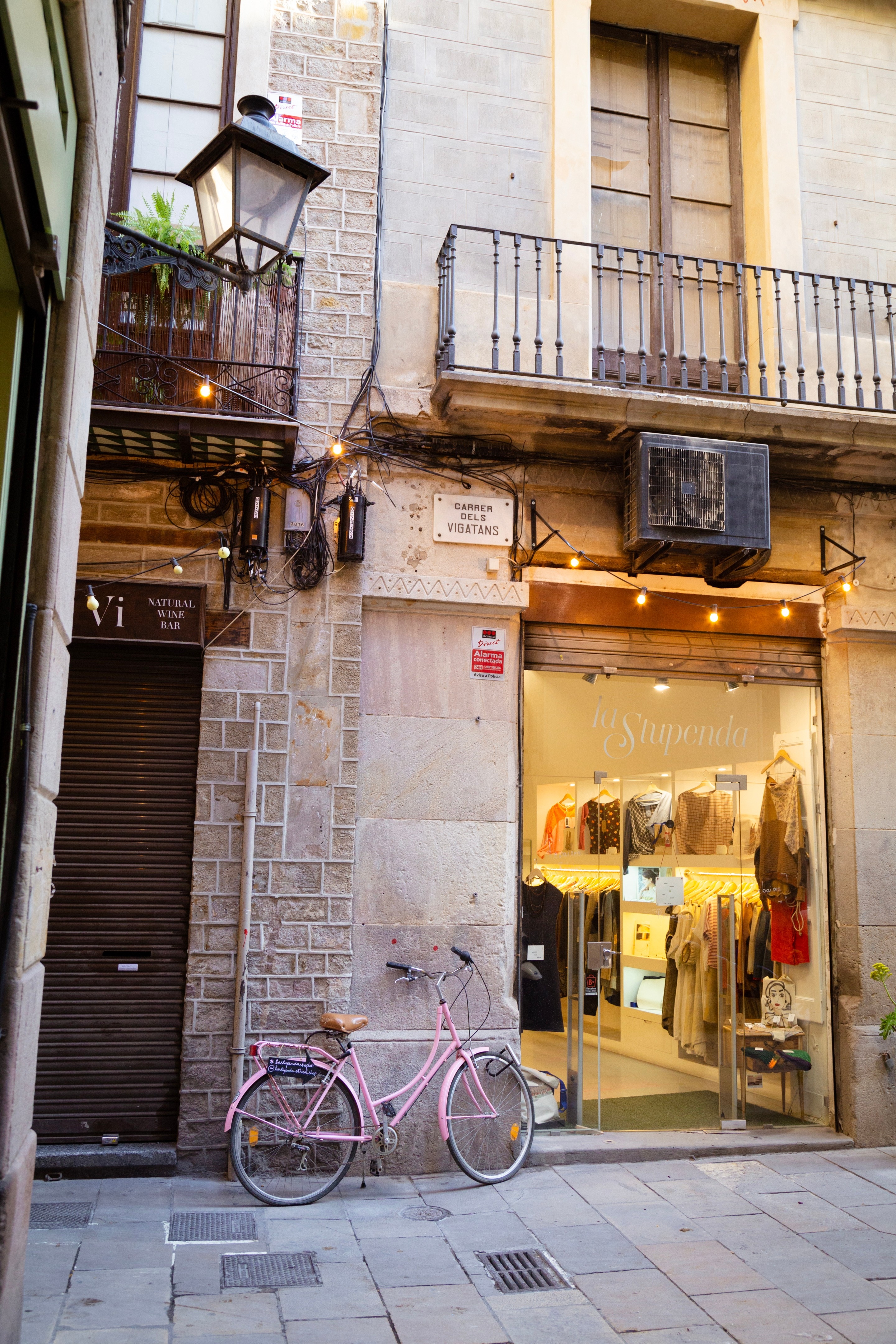 A pastel pink colour bike leaning against the wall next to an inviting clothes shop on the streets of Barcelona.