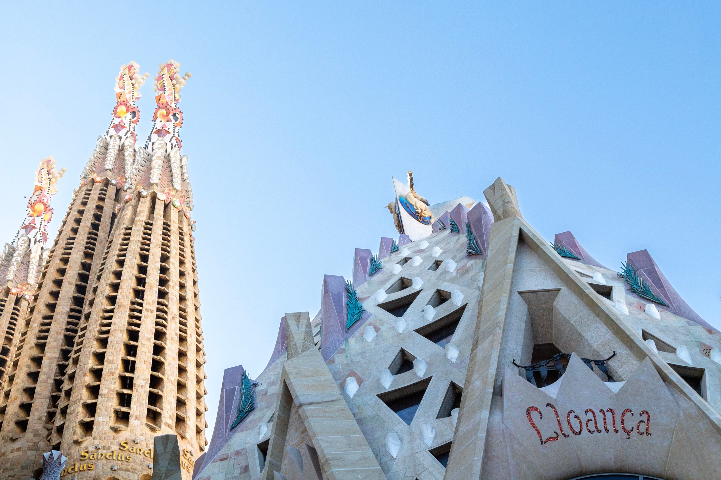 The word 'lloanica' meaning 'praise' inscribed onto the foreground building with three of the intricate tile covered spires on La Sagrada Familia in the background