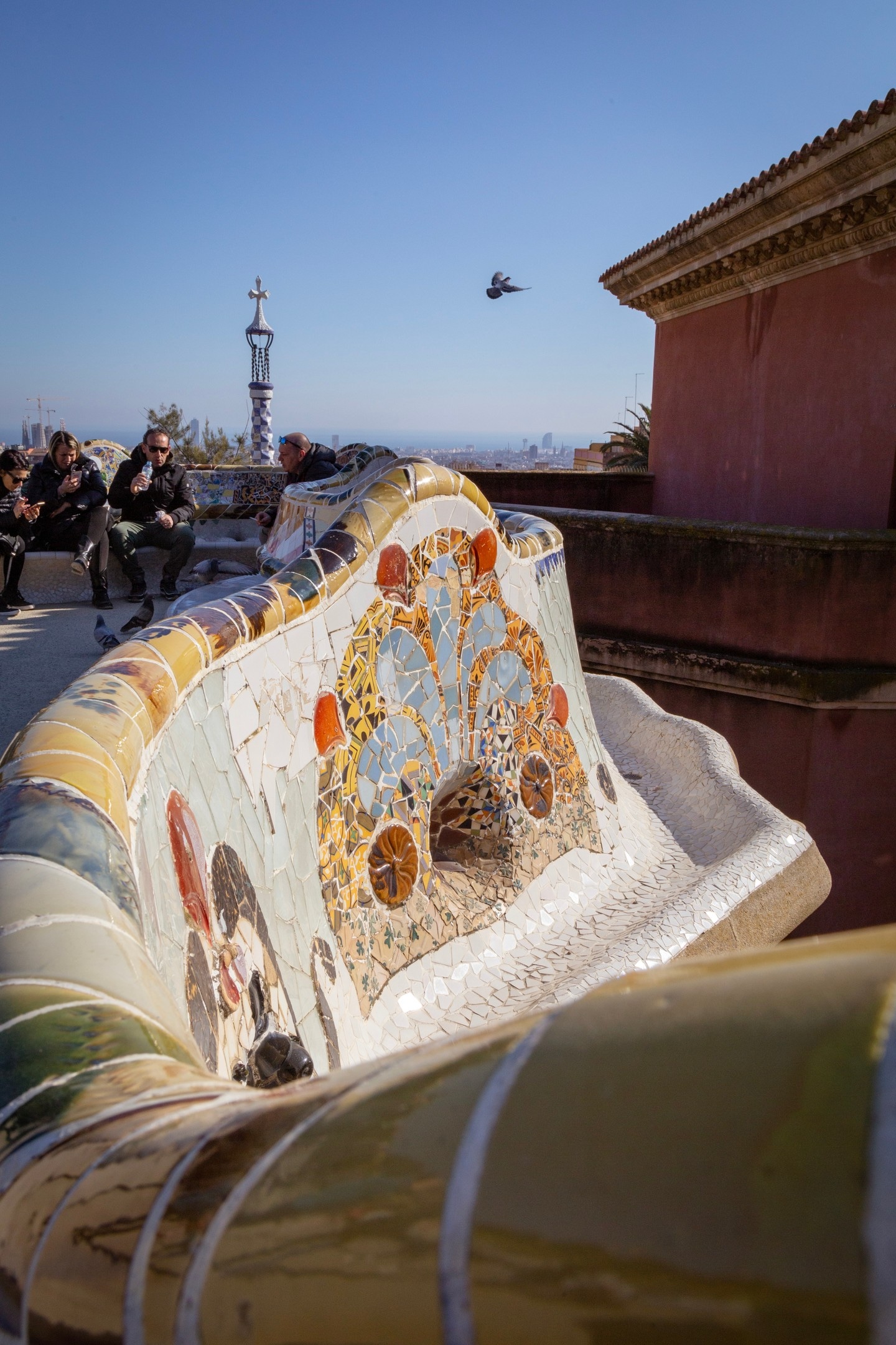 Tiled entrance tower in the background, with a pigeon caught mid-flight and the tiled seating area in the foreground, at Parc Guell, Barcelona.