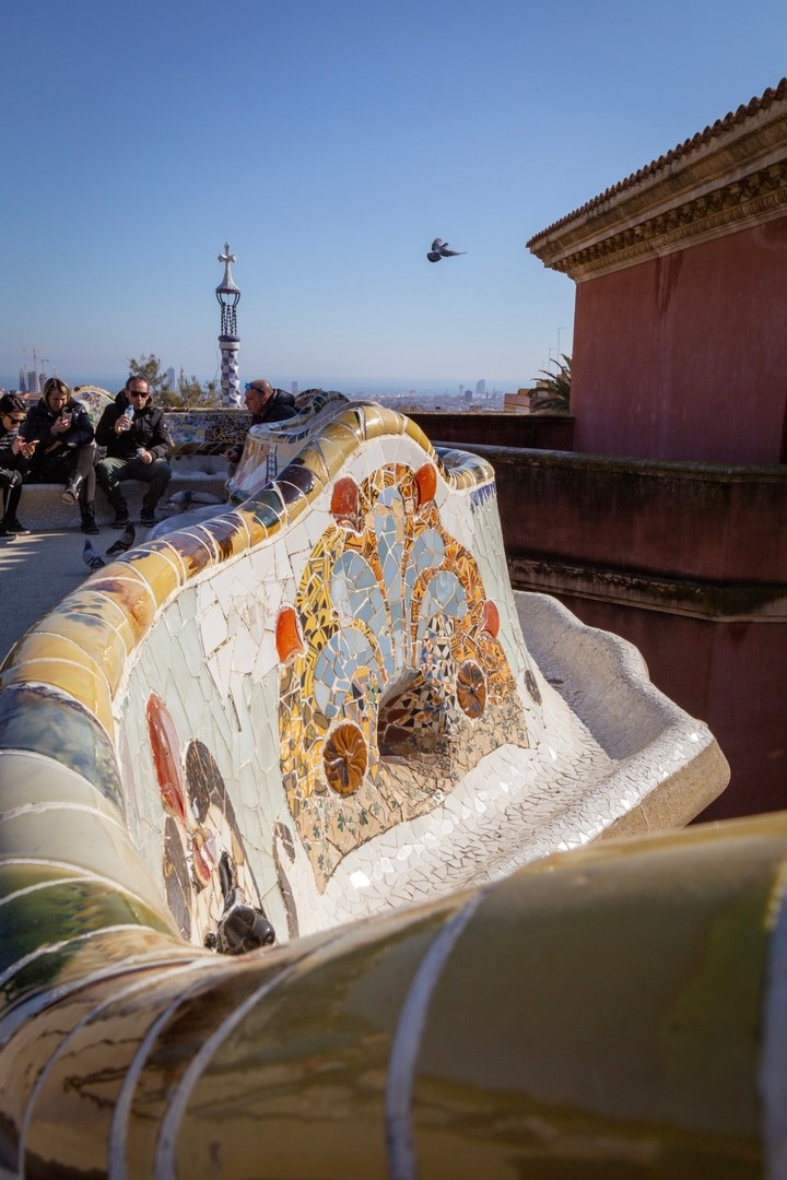 Tiled entrance tower in the background, with a pigeon caught mid-flight and the tiled seating area in the foreground, at Parc Guell, Barcelona.
