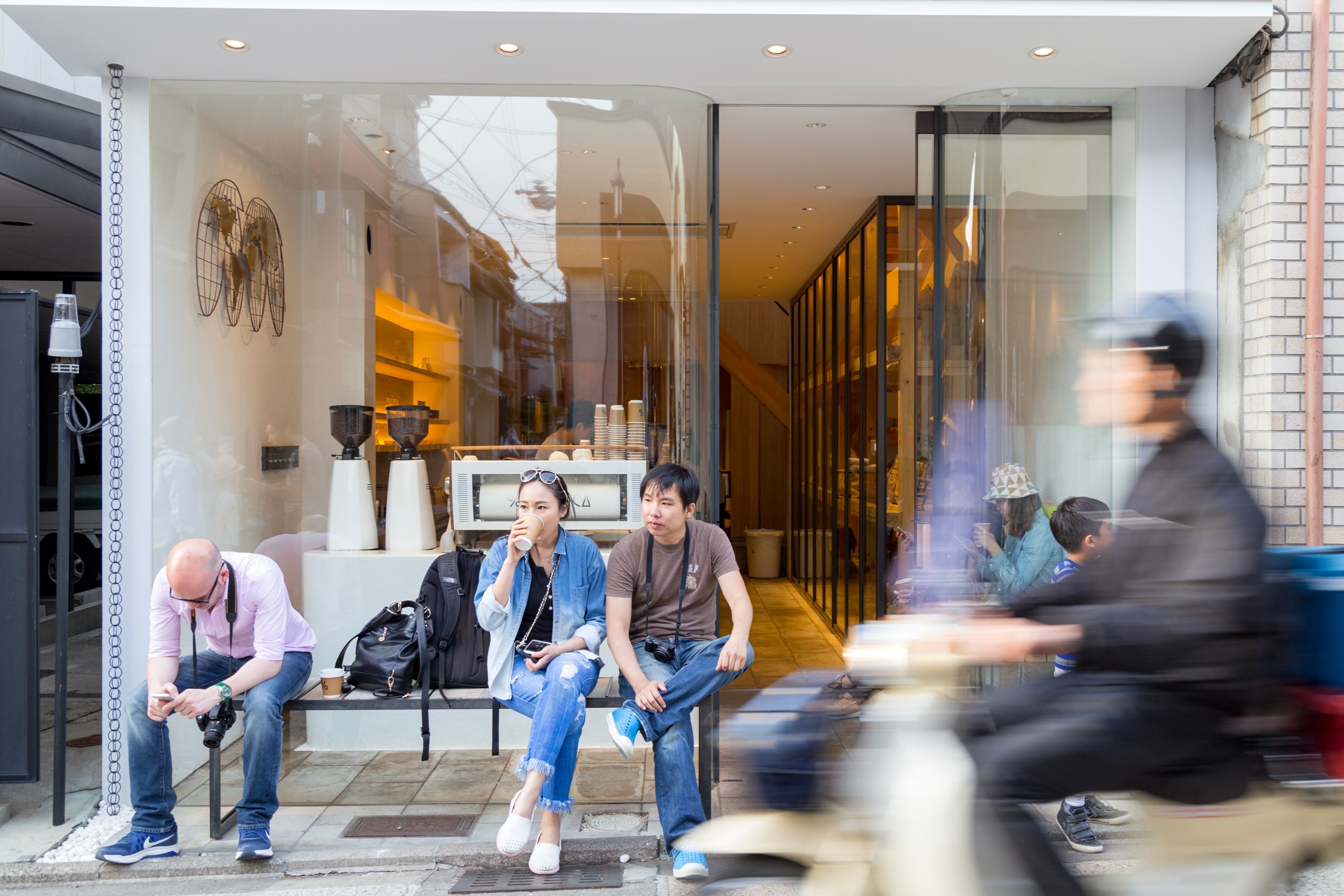 Tourists relaxing outside AKR, a trendy cafe in Kyoto. A man on a moped enters the scene from the right, blurred and adding a sense of movement to an otherwise tranquil scene — Kyoto, Japan