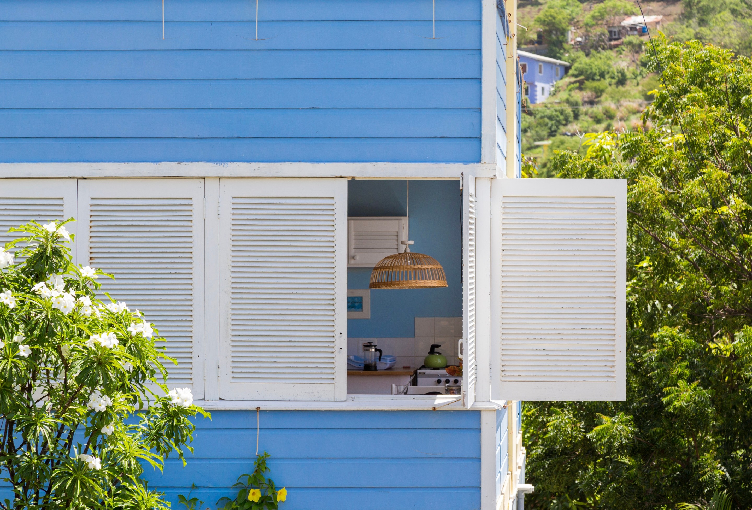 Looking through an open white shutter, a wicker ceiling lampshade can be seen hanging within a simple kitchen, it's inner blue walls matching with wooden slatted facade of the property, creating a tranquil Caribbean scene — Saint Vincent and the Grenadines