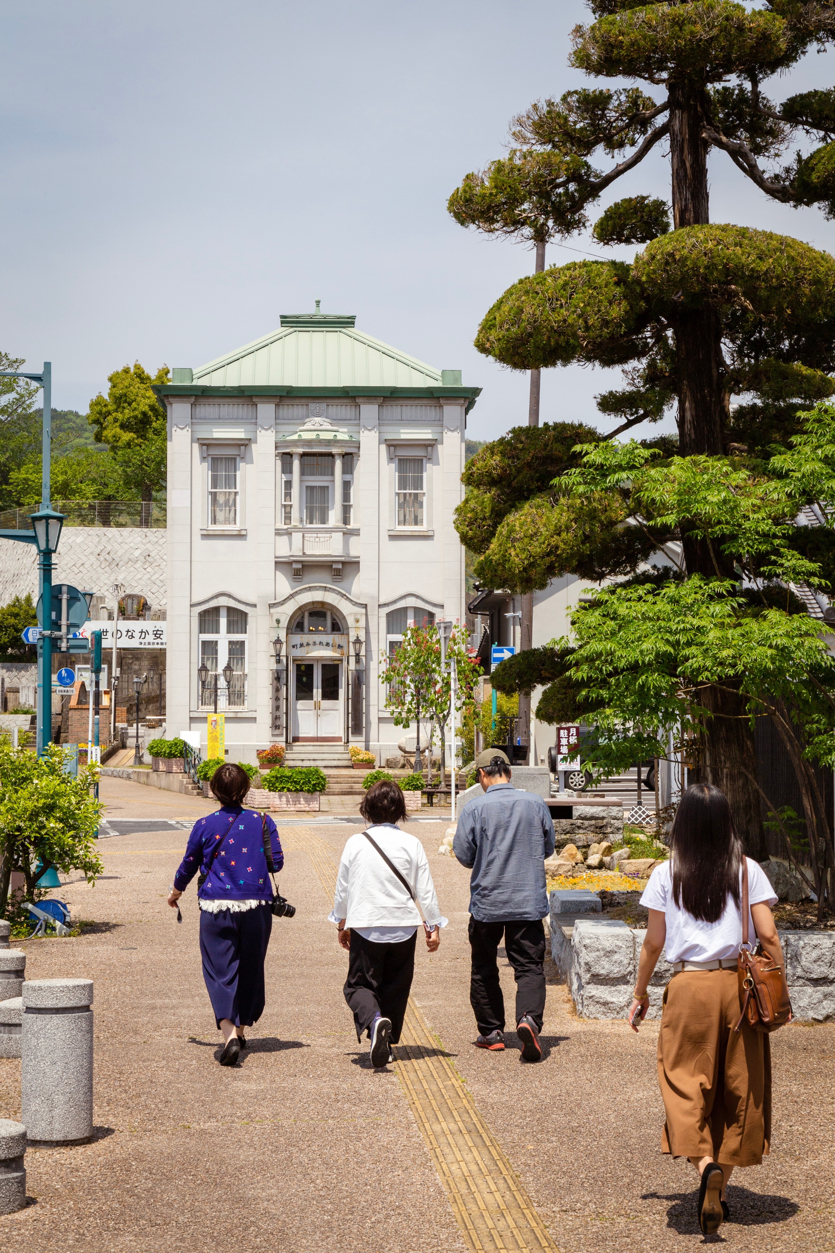 People walking down a road in Yanai, with an old bank building ahead of them and shaped trees to the side — Yanai, Yamaguchi Prefecture, Japan