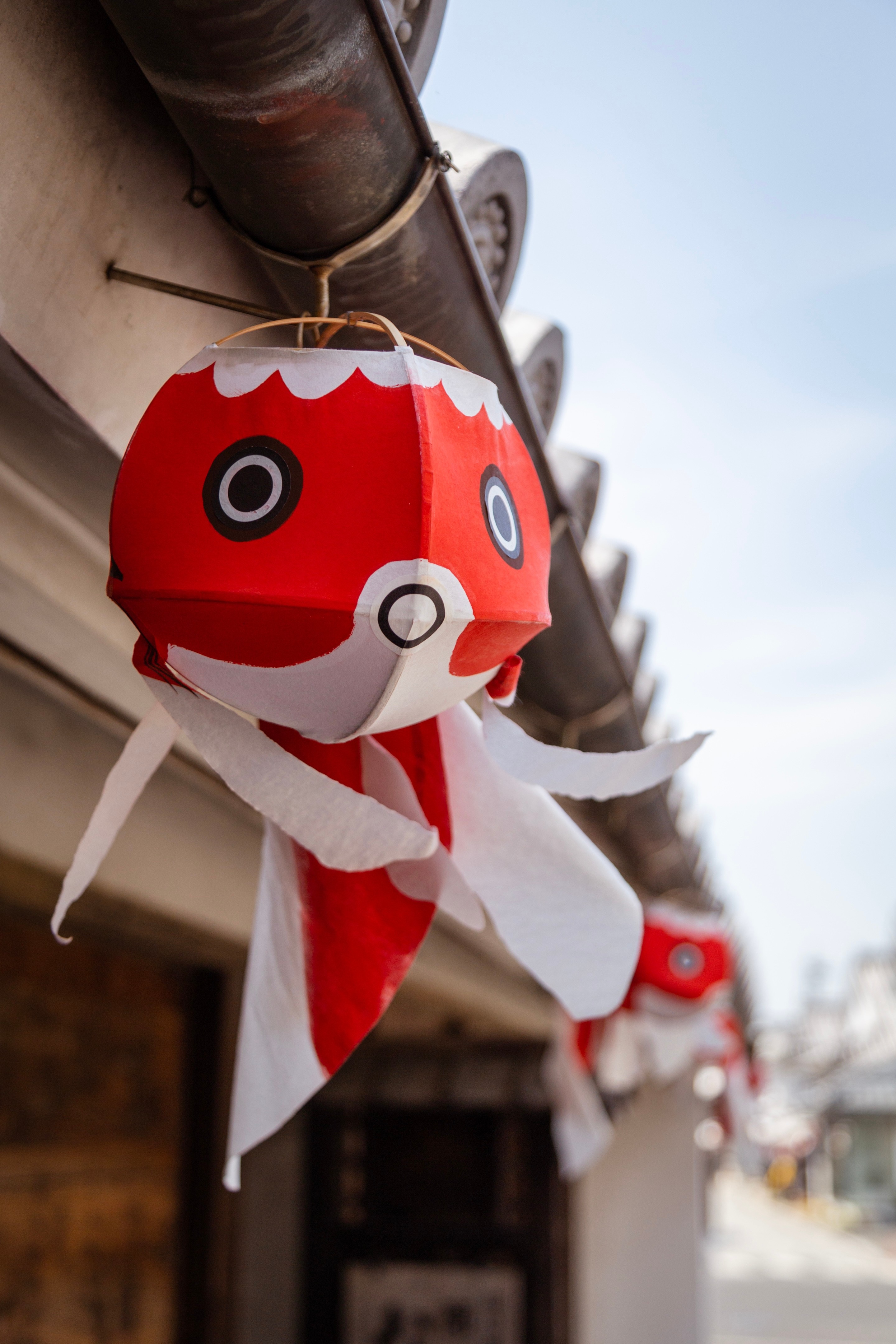 A single paper goldfish lantern hangs from a metal gutter with more blurred lanterns in the background — Yanai, Yamaguchi Prefecture, Japan