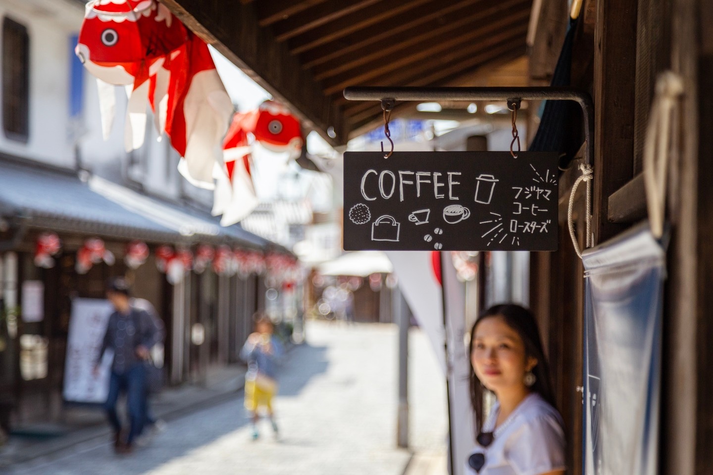 A shop sign in focus in the foreground, with 'coffee' written along with some simple coffee illustrations. In the blurred background is a smiling woman standing the in the shop entrance surrounded by paper goldfish lanterns — Yanai, Yamaguchi Prefecture, Japan