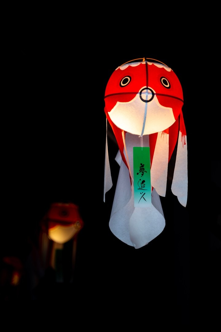 A single illuminated paper goldfish lantern suspended in the air against a black background, more blurred lanterns can vaguely be seen in the background — Yanai, Yamaguchi Prefecture, Japan