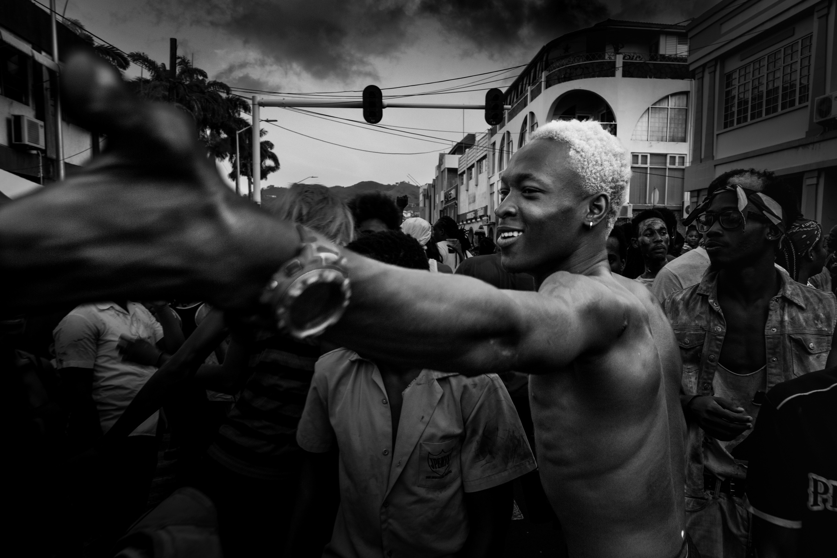 Black and white street party scene. A man with bleached white hair smiles whilst welcoming a friend. Taken at Vincy Mas, the annual carnival — Kingstown, Saint Vincent and the Grenadines