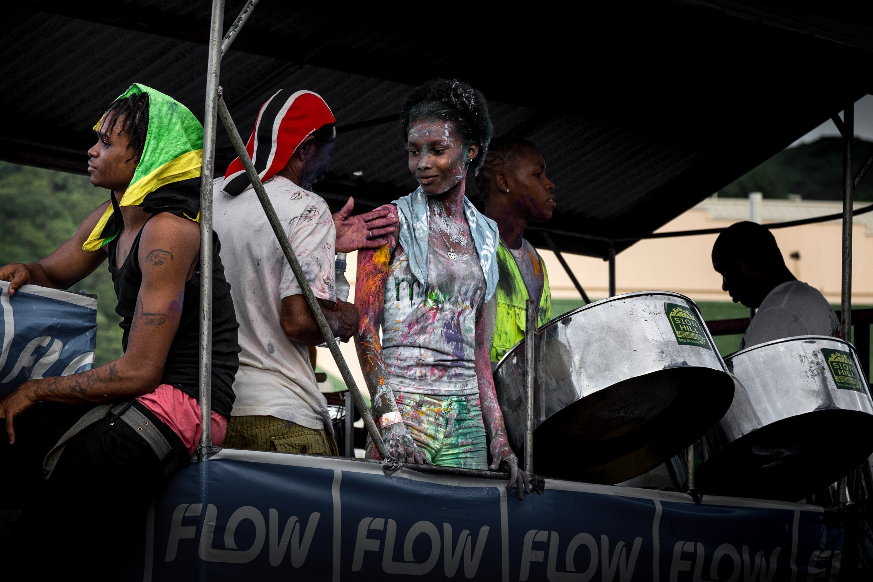 Party revellers covered in vibrant paints, seen talking to each other, whilst a drummer plays the steel drums next to them. Taken at Vincy Mas, the annual carnival — Kingstown, Saint Vincent and the Grenadines