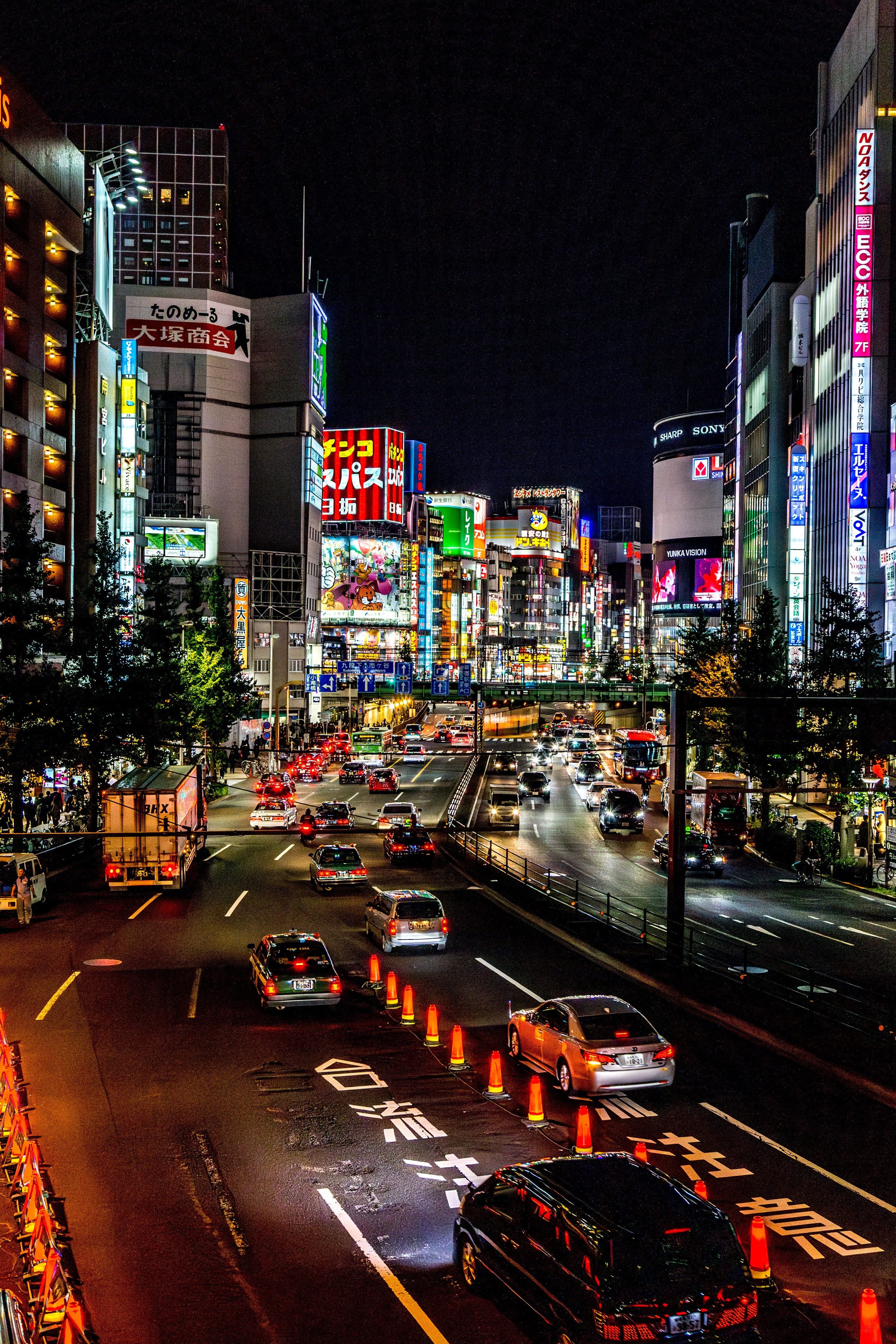 A night time shot of a busy road in Nishi-Shinjuku, with the bright neon illuminated lights and signs leading the eye into the cityscape beyond — Nishi-Shinjuku, Tokyo, Japan