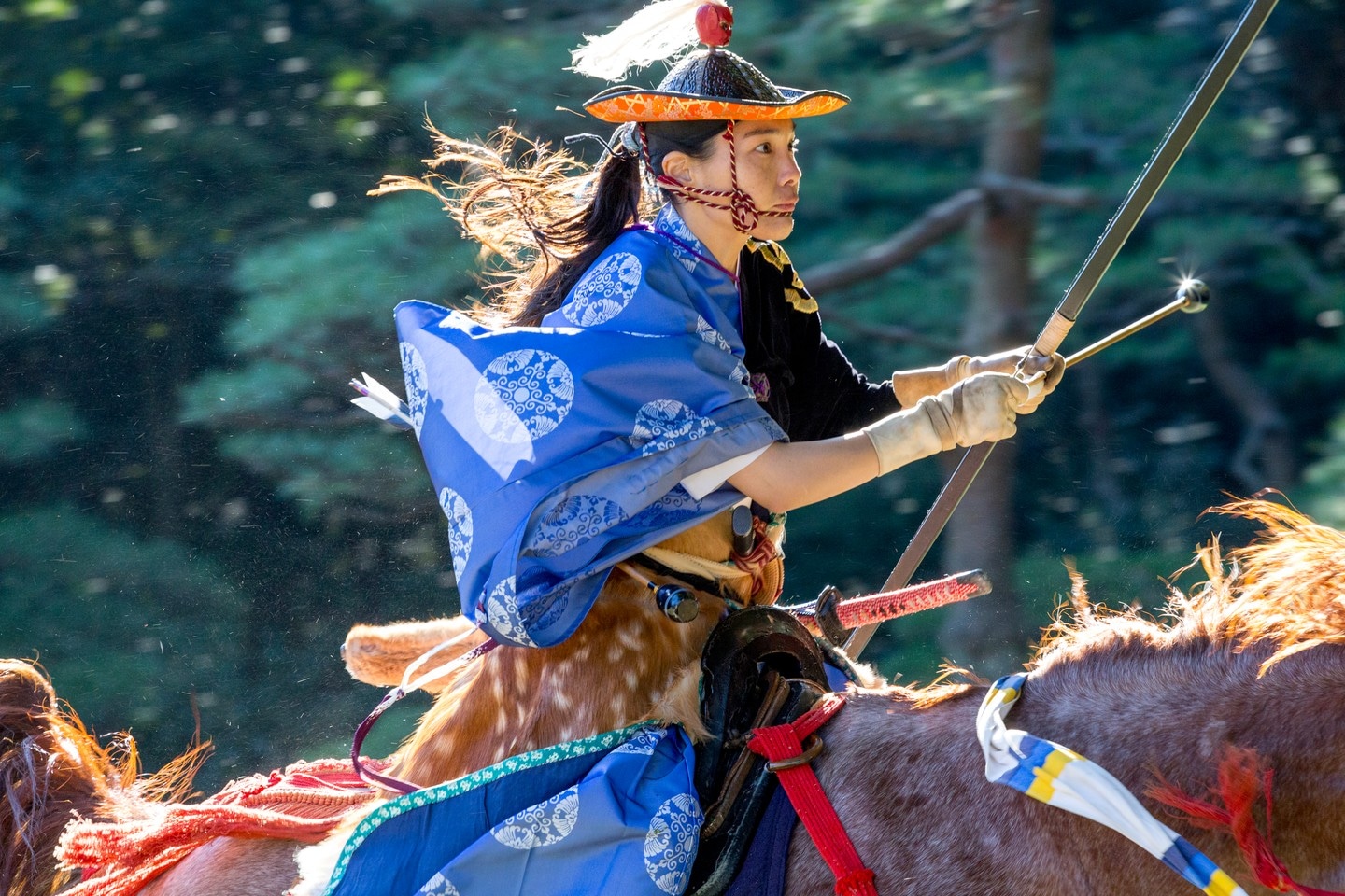 An uber cool female archer in traditional blue dress rides a horse, whilst readying her bow to release at her target, in this scene taken from an archery contest in Yoyogi Goen (park) — Marunouchi, Tokyo, Japan