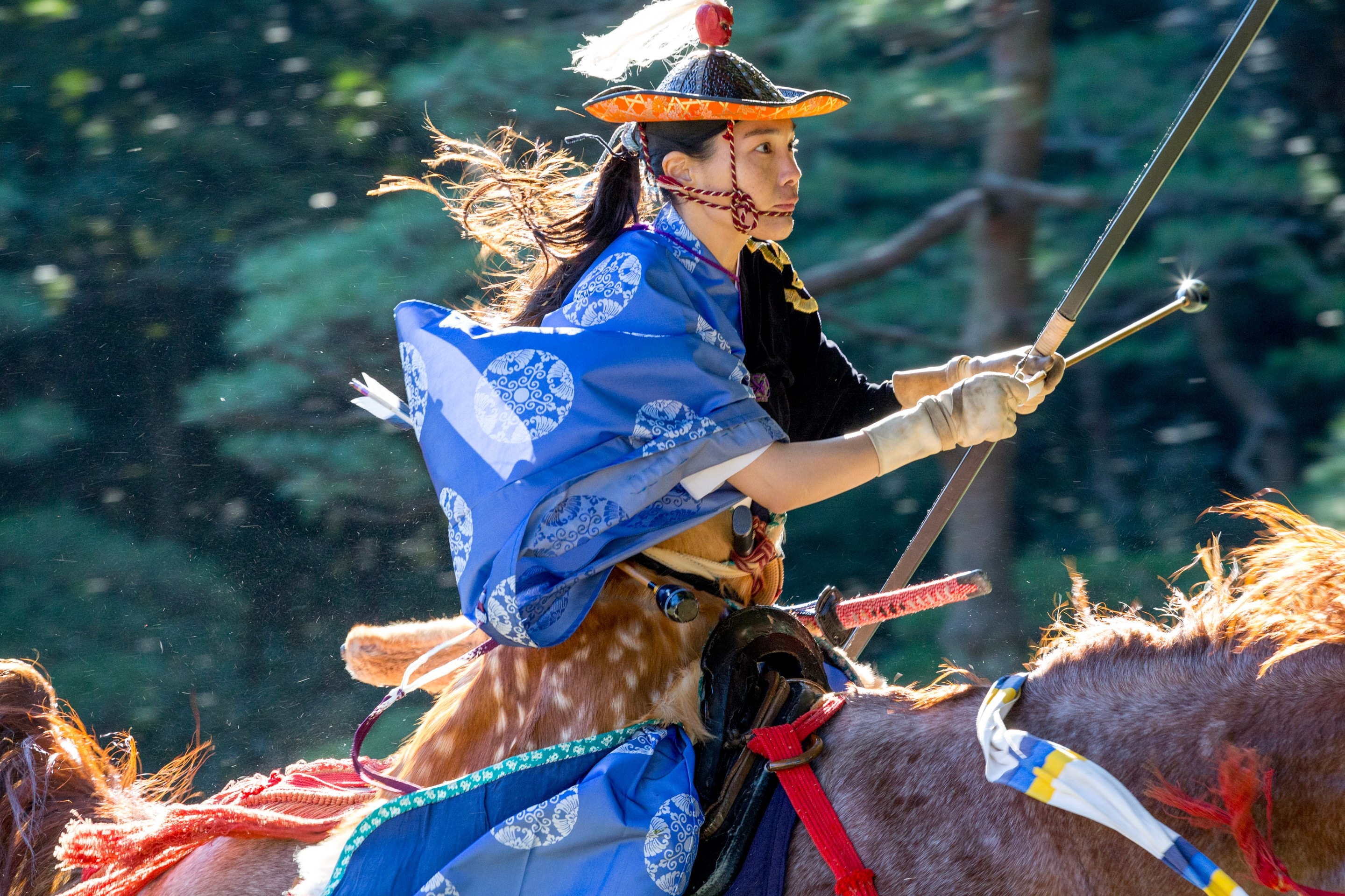 An uber cool female archer in traditional blue dress rides a horse, whilst readying her bow to release at her target, in this scene taken from an archery contest in Yoyogi Goen (park) — Marunouchi, Tokyo, Japan