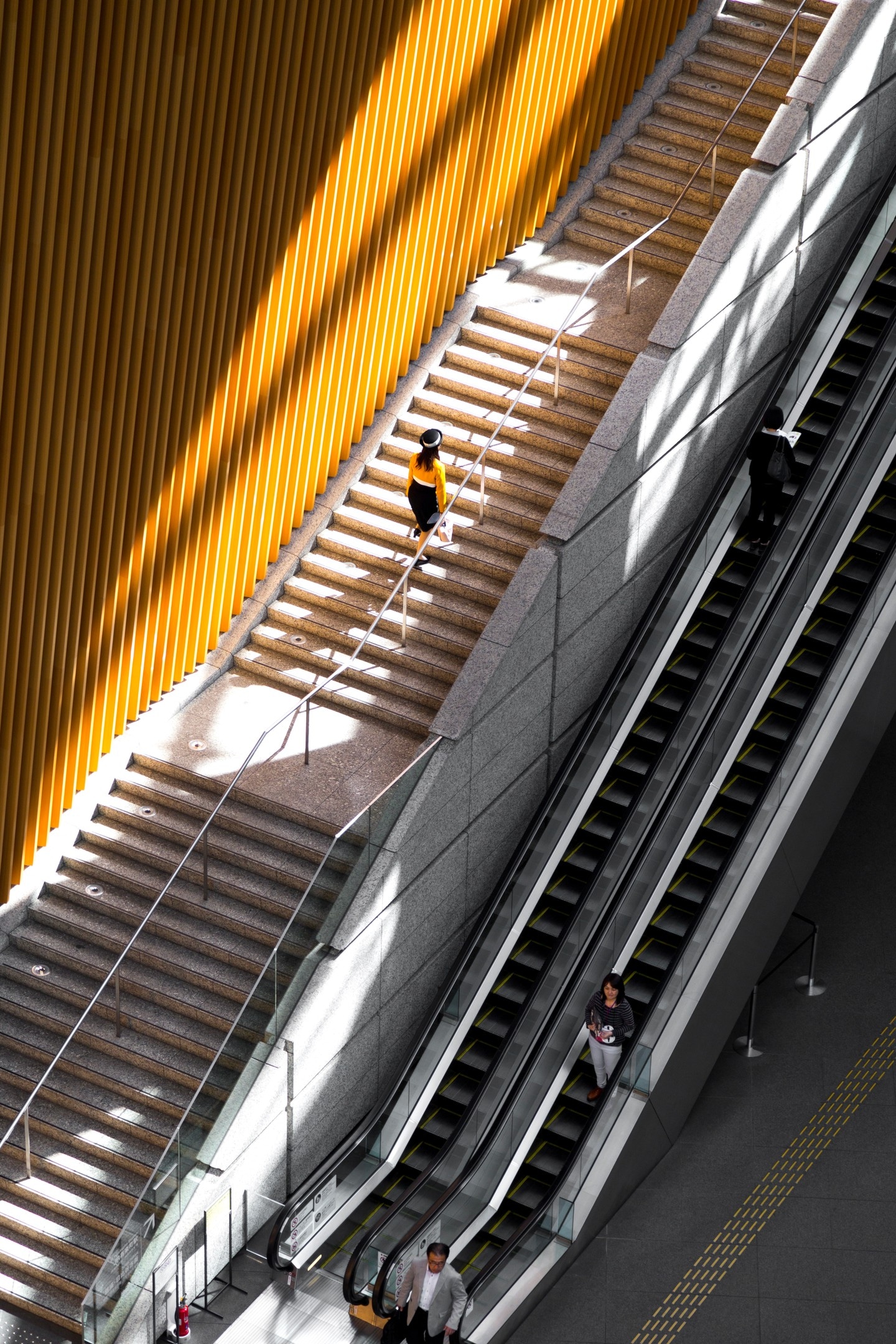 A woman in an orange top walks up a long concrete staircase, itself lined with matching orange walls. The sun both illuminates and casts long shadows creating striking contrast and interest. Taken at the Tokyo International Forum building — Marunouchi, Tokyo, Japan